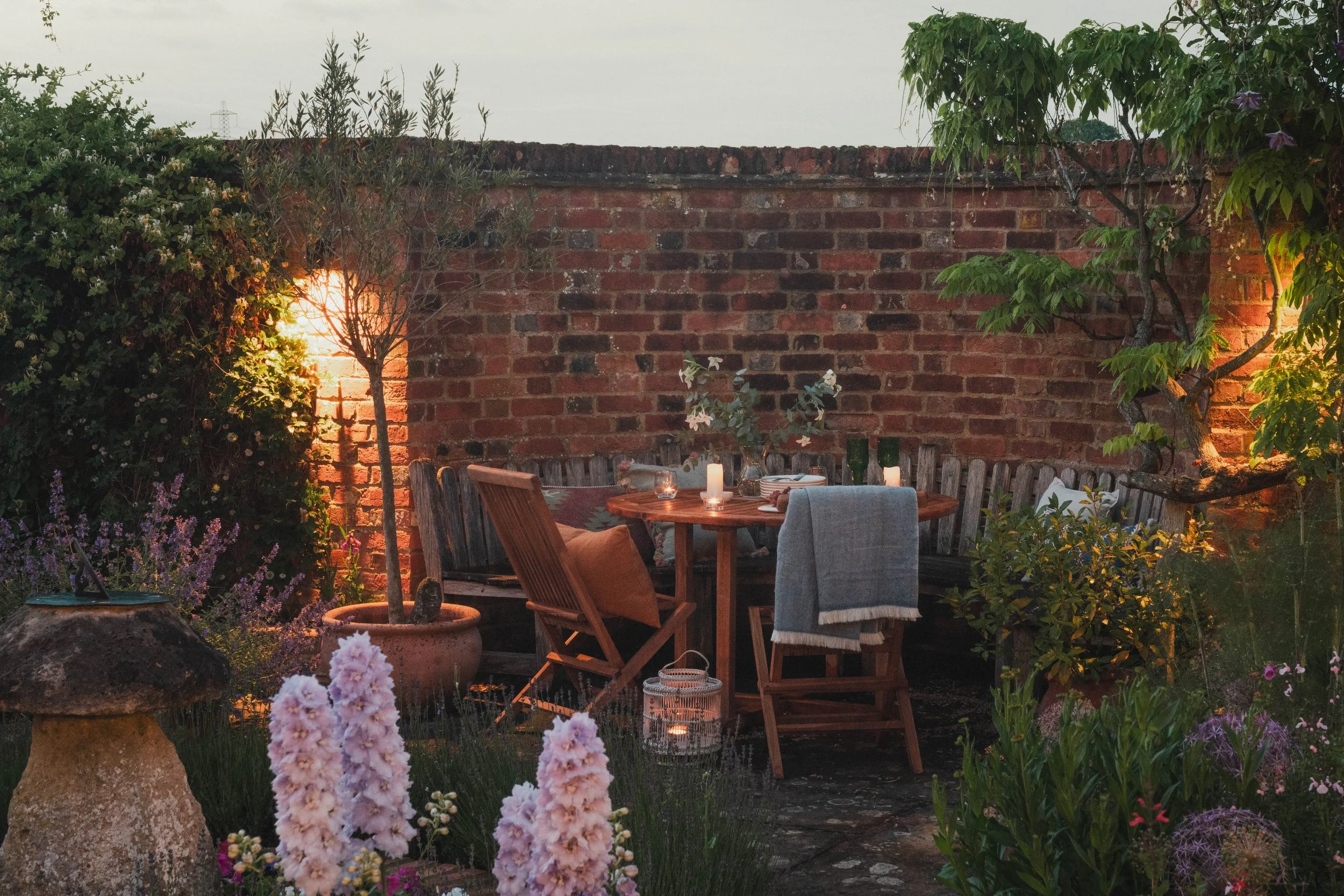 A cozy outdoor garden dinner setup with a round wooden table, chairs with cushions, candles, and a brick wall surrounded by lush flowers and greenery.