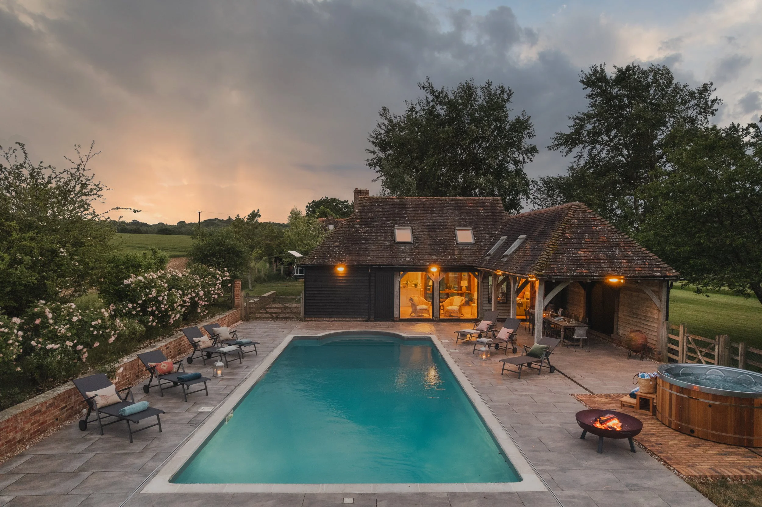 A backyard with a swimming pool, surrounded by lounge chairs and a brick patio. There is a house with large windows and outdoor lighting, a hot tub, and lush green trees and shrubs around. The sky is cloudy with a setting sun in the background.