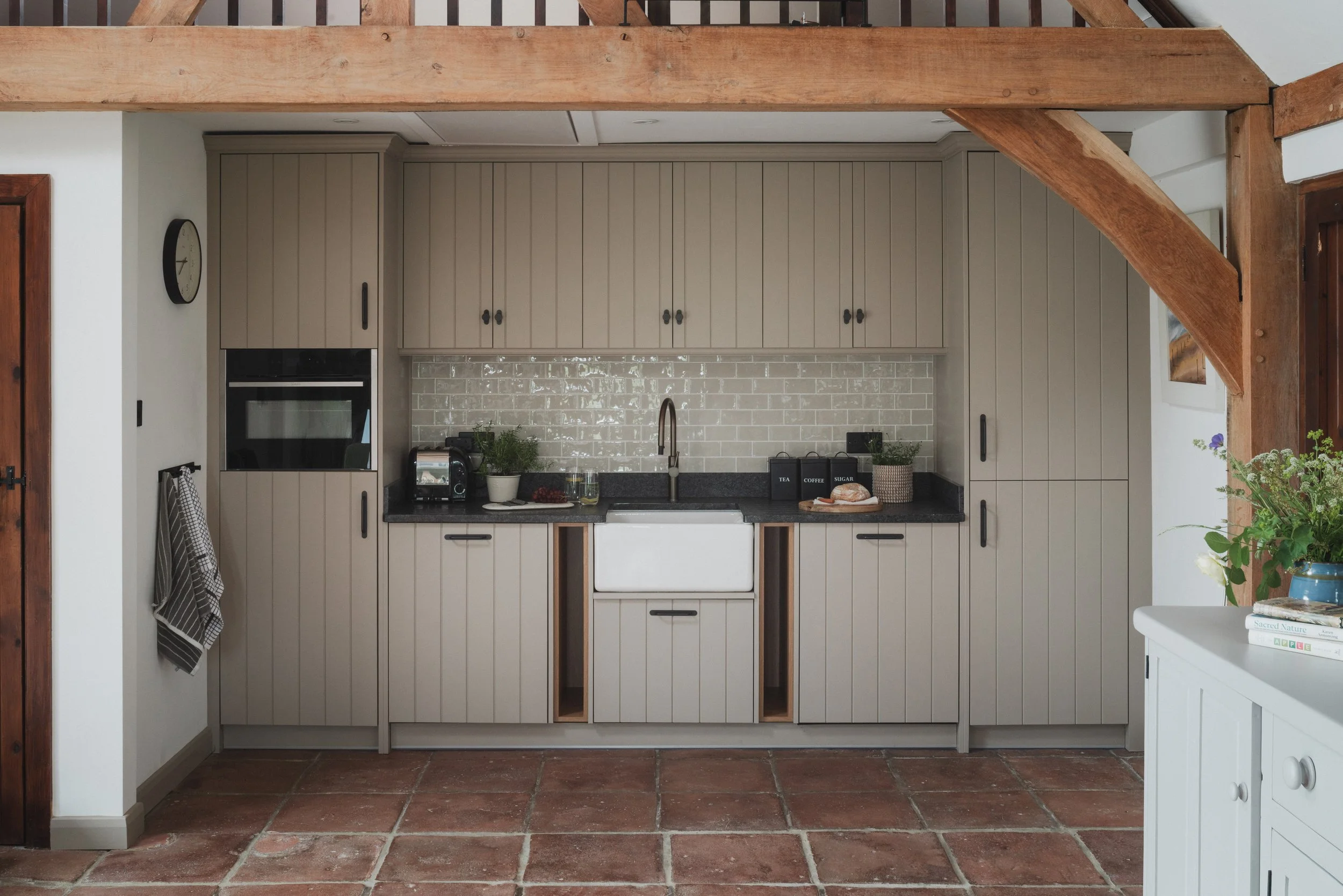 Kitchen with beige cabinets, black countertops, white sink, brick backsplash, and wooden beams overhead.
