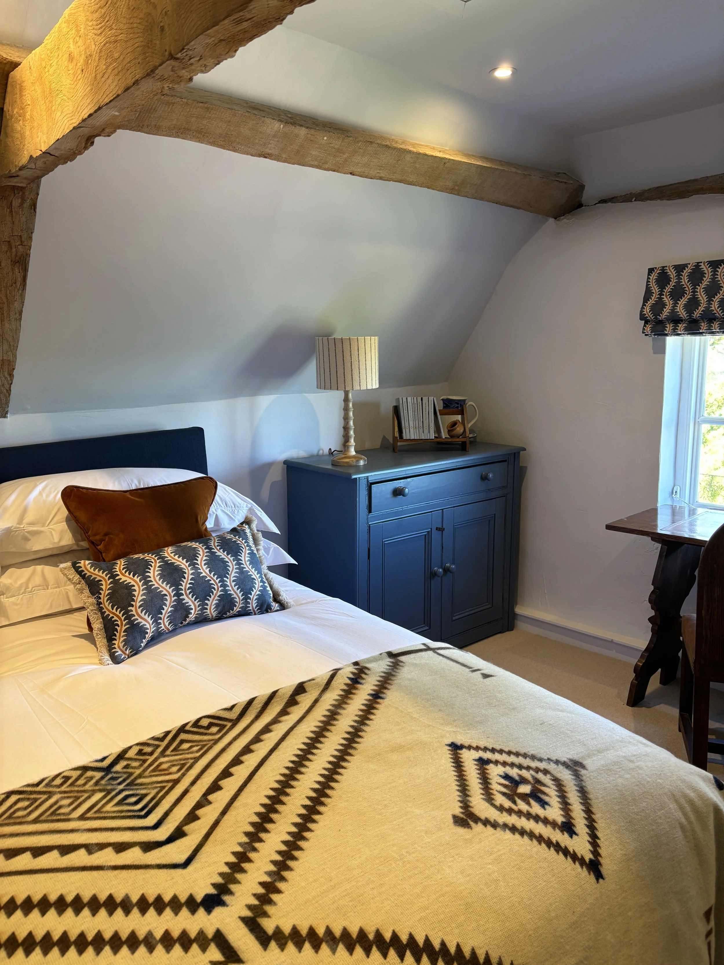Cozy bedroom with a bed covered in a geometric patterned blanket, a brown pillow, and a decorative pillow. A blue dresser holds a table lamp, books, and decorative items. A window with a patterned valance lets in natural light. Exposed wooden beams a