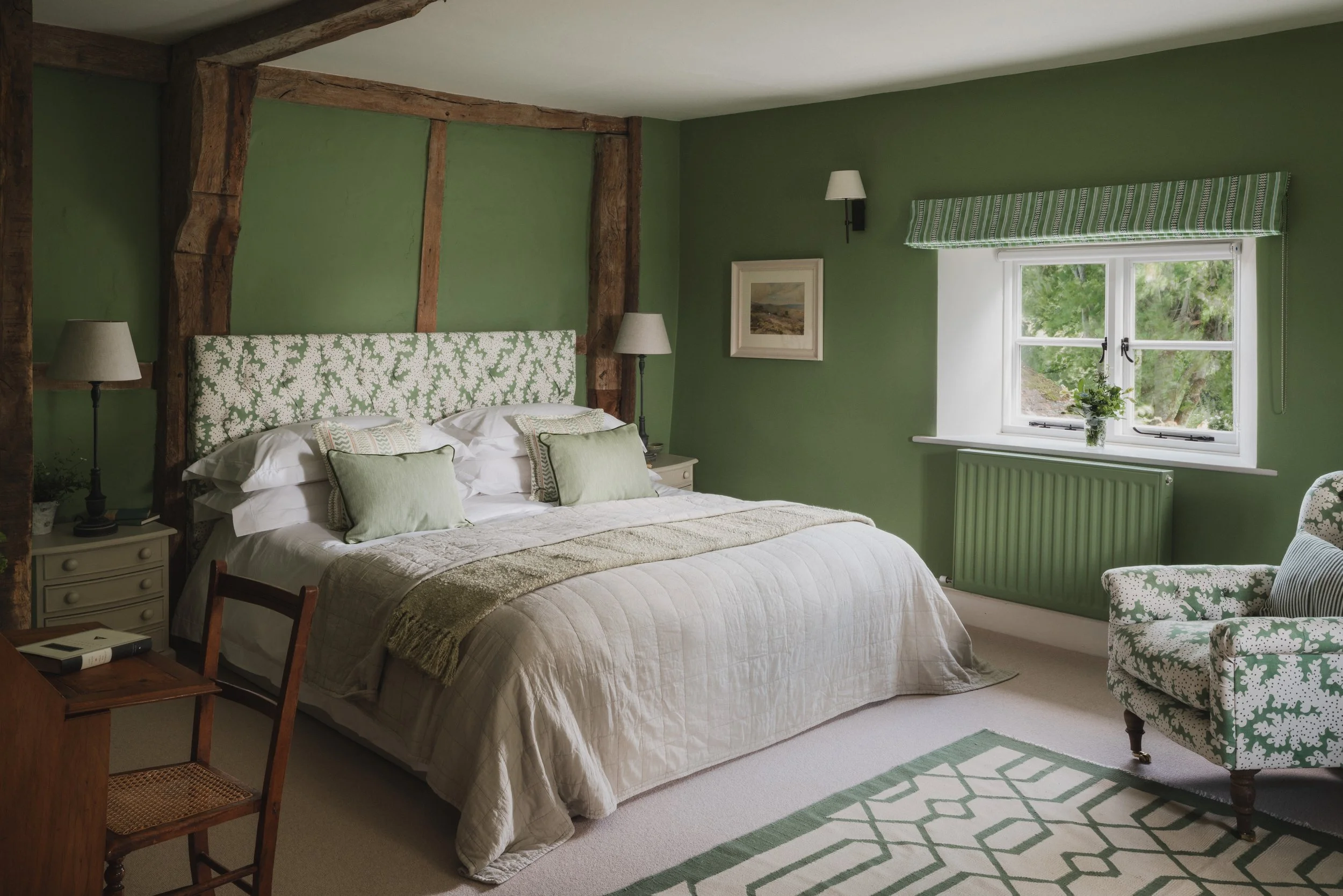 A cozy bedroom with green walls, a bed with a patterned headboard, multiple pillows, and a neutral bedspread. There is a window with a green-striped valance and a plant on the sill, a framed landscape picture on the wall, a pair of lamps on nightstan