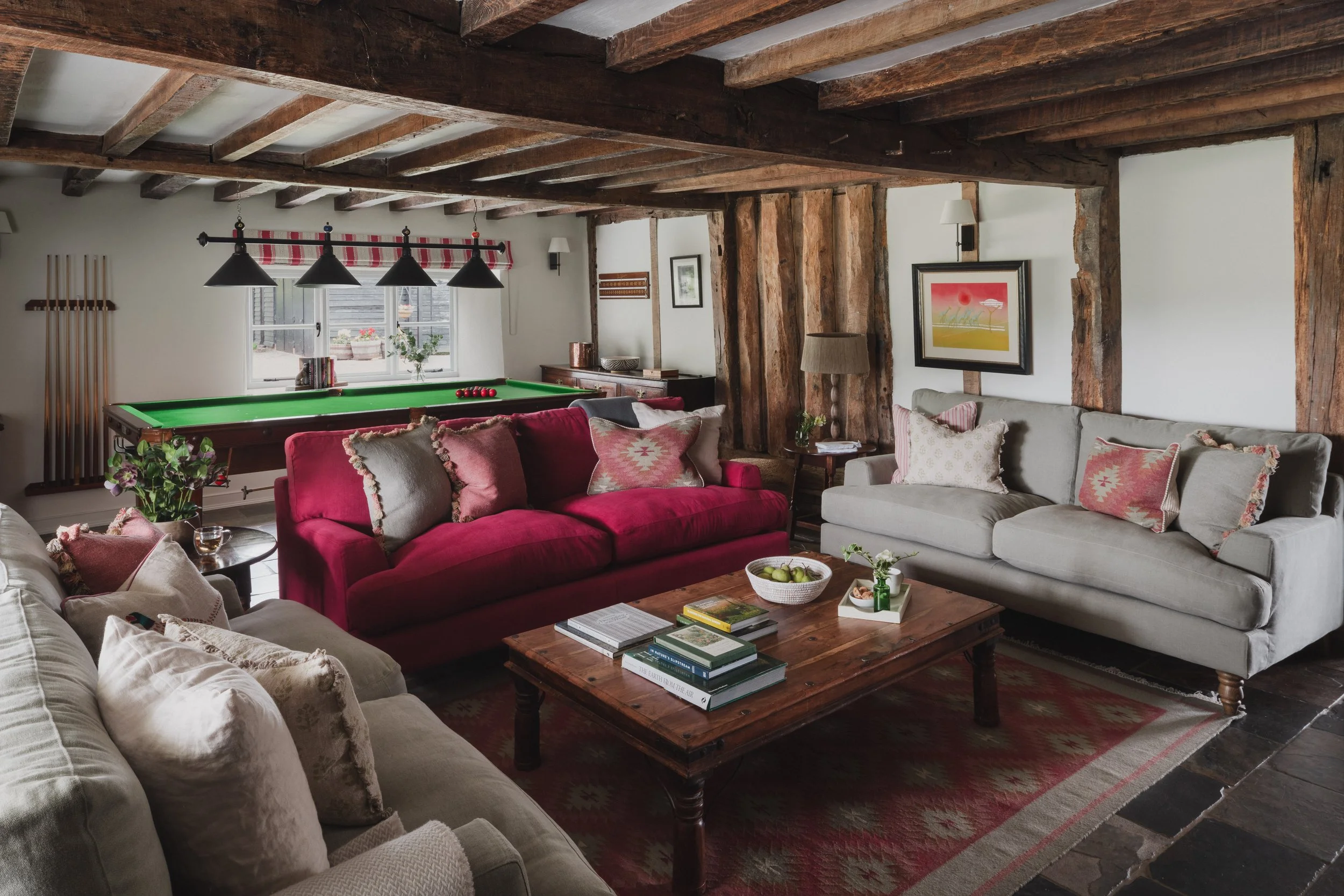 Living room featuring exposed wooden ceiling beams, a red sofa, a beige sofa, a wooden coffee table with books and a tray, a pool table by the window, framed artwork on white walls, and various lamps and decorative items.