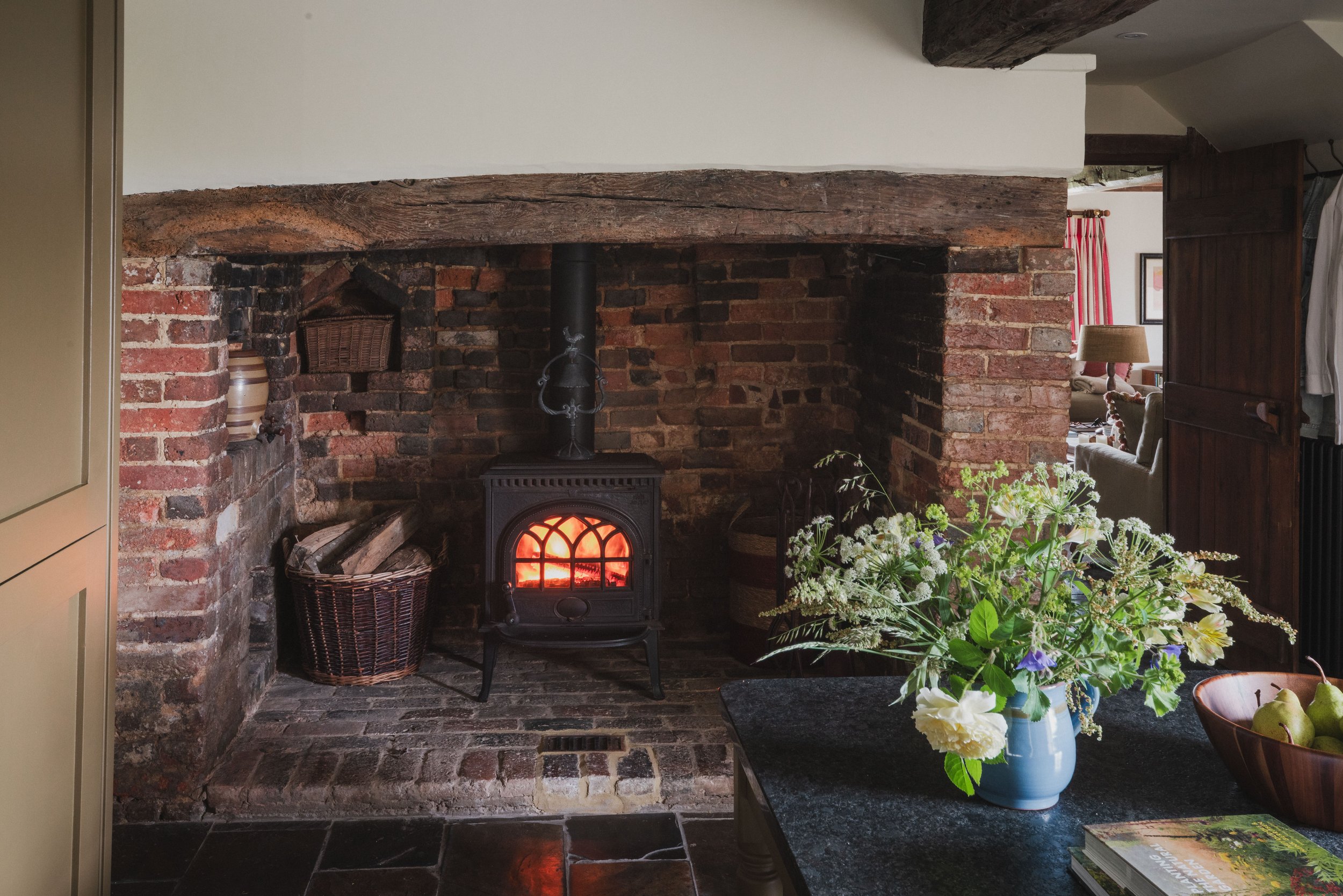 A cozy living room corner with a wood-burning stove set within a brick fireplace, a basket of firewood, a vase of flowers, and a pot of pears, with part of a seating area visible in the background.