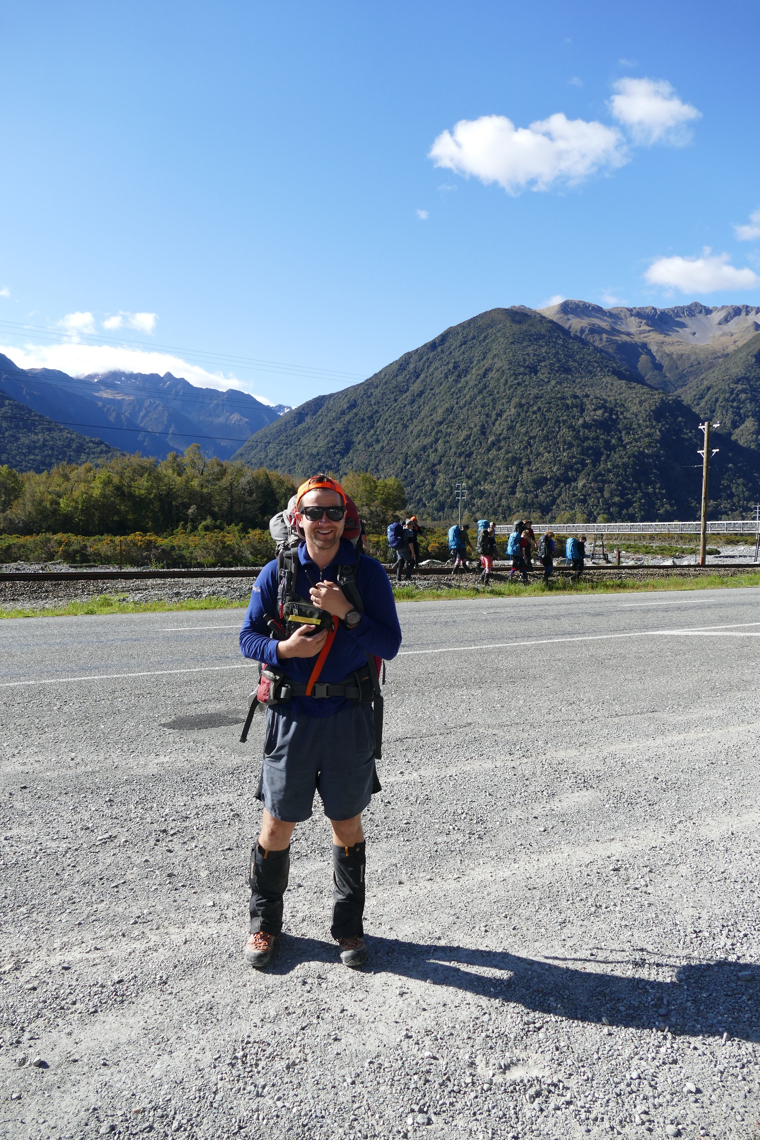 A man dressed in outdoor hiking gear standing by a gravel roadside, with a group of hikers walking along the train tracks behind him and a mountain landscape with green hills and a blue sky in the background.