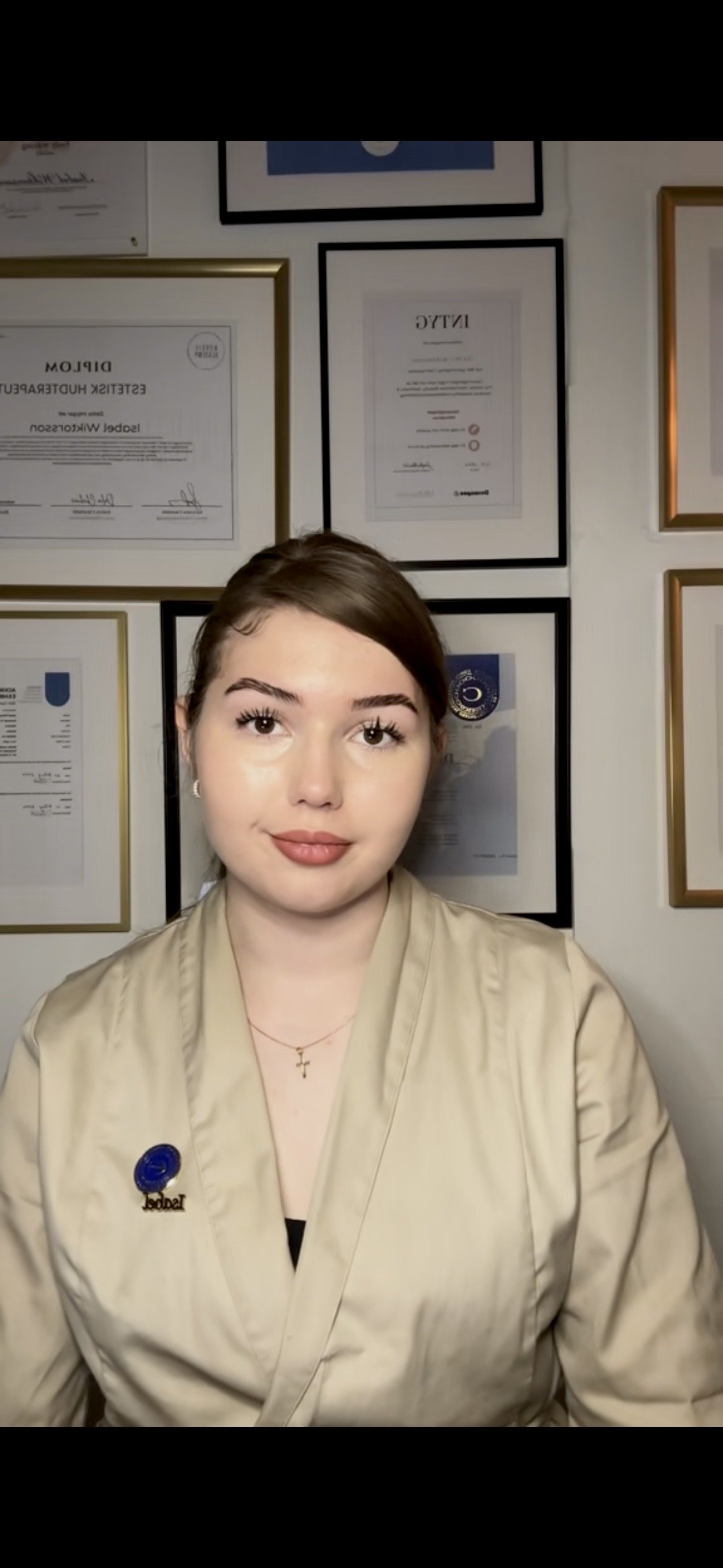 En ung kvinna med brunt hår och ljus hud, klädd i en beige uniform, står framför en vägg fylld med certifikat och diplom.