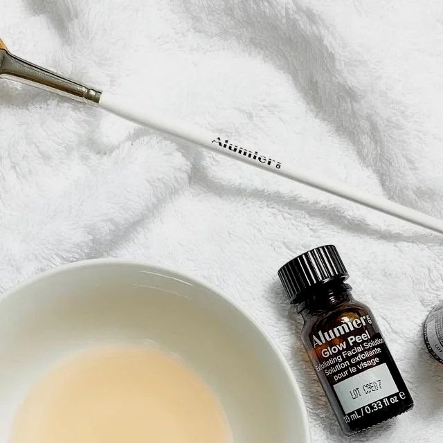 En vit flaska av märket Alumier med etiketten Glow Peel ligger på ett vitt, fluffigt material tillsammans med en vit skål och en nagelpensel.