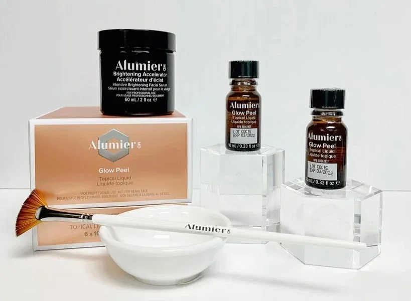 Skönhetsprodukter inkluderande en burk med Alumier Brightening Accelerator, två små flaskor med Alumier Glow Peel, en handborste, en vit skål och ett kosmetiskt box med Alumier emblem.