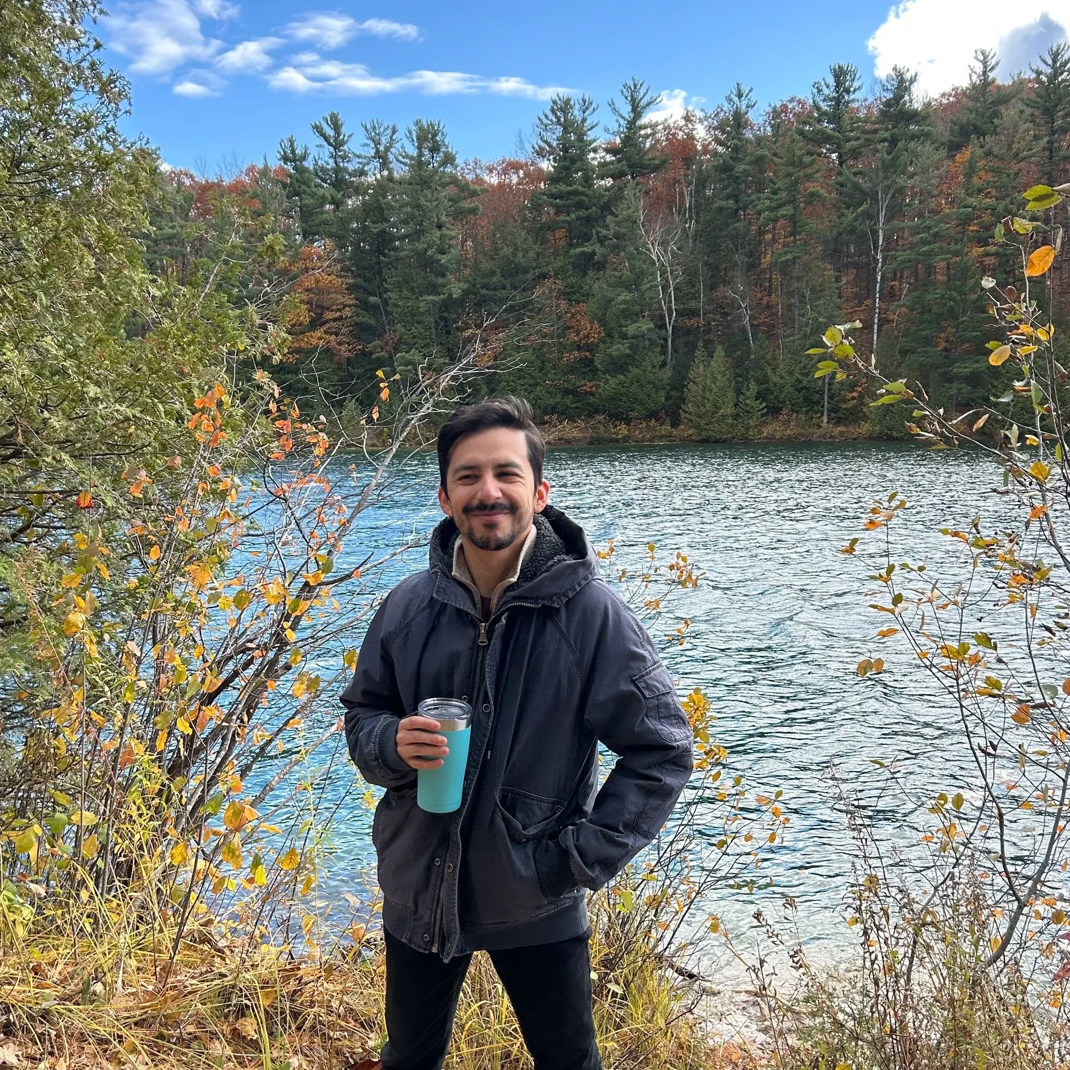 A man smiling outdoors near a body of water, holding a blue and white insulated mug, with autumn-colored trees and a partly cloudy sky in the background.