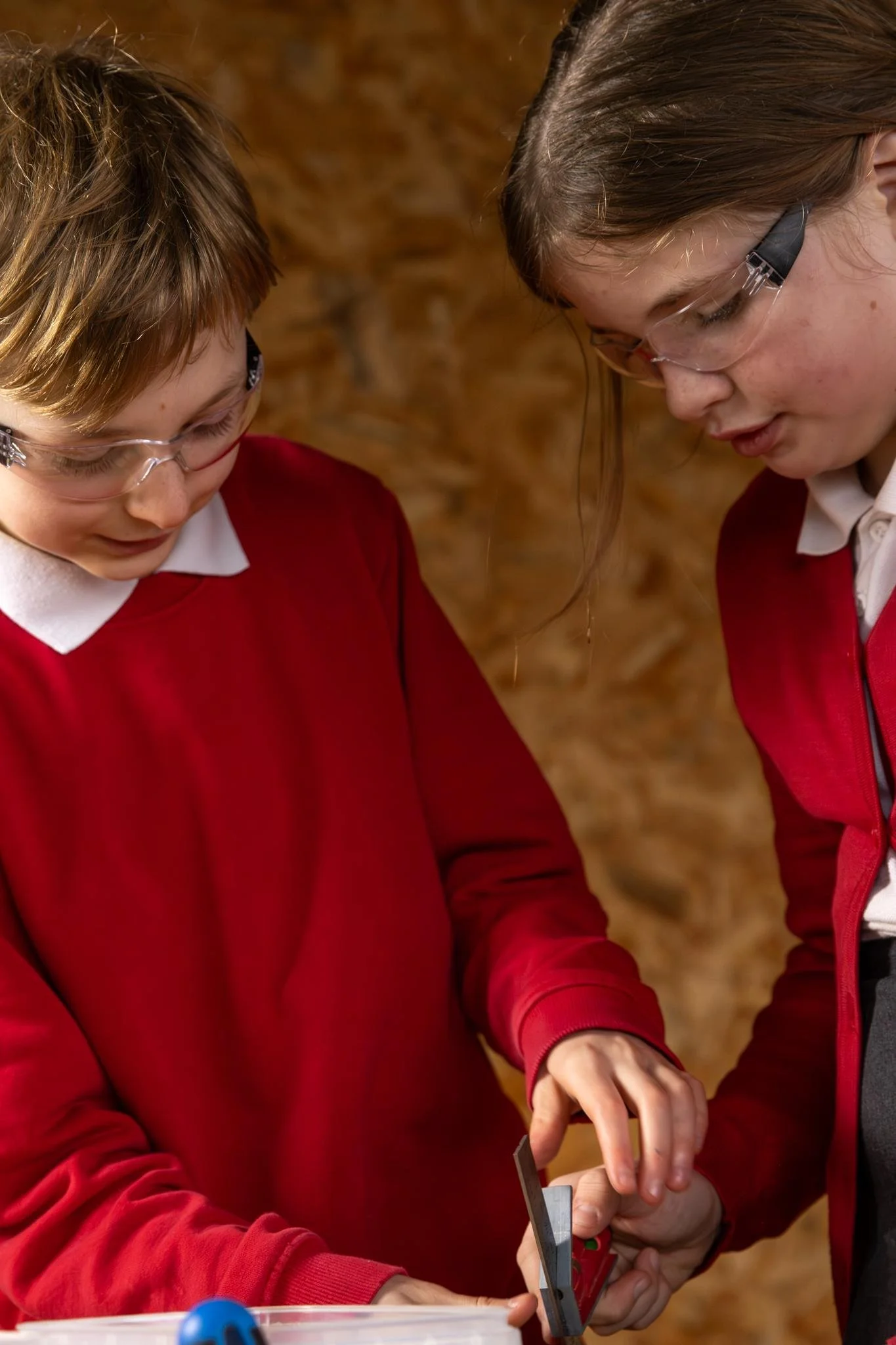 Two children wearing safety goggles and red school uniforms are working together on a carpentry project.