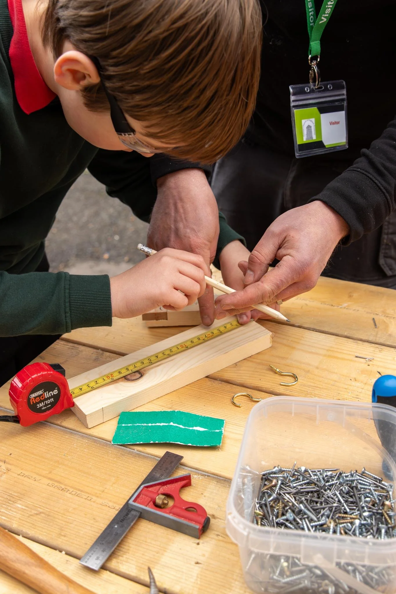 People conducting woodworking activity with various tools and materials on a wooden table including screws, a measuring tape, rulers, a saw, and wood pieces.