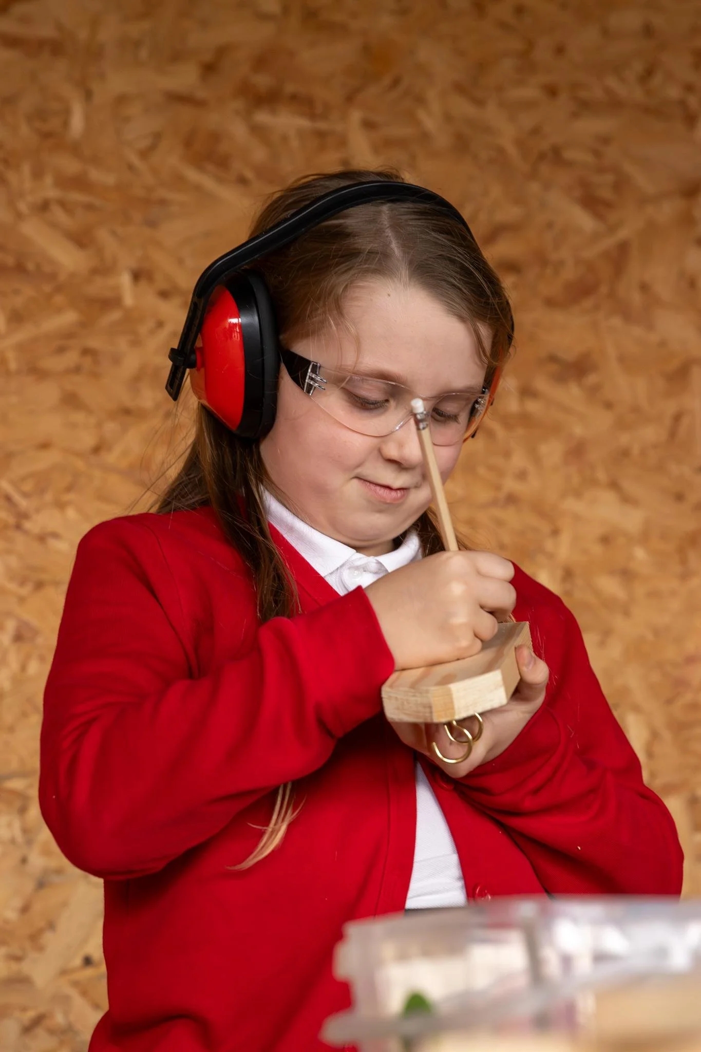 A young girl in a red jacket and glasses, wearing safety ear protection, is holding a piece of wood object and a pencil.