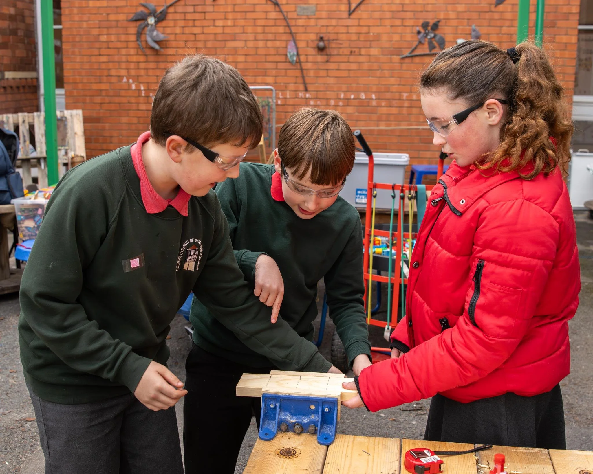Three children wearing safety goggles work together on a woodworking project outdoors, with a brick wall and various garden tools in the background.