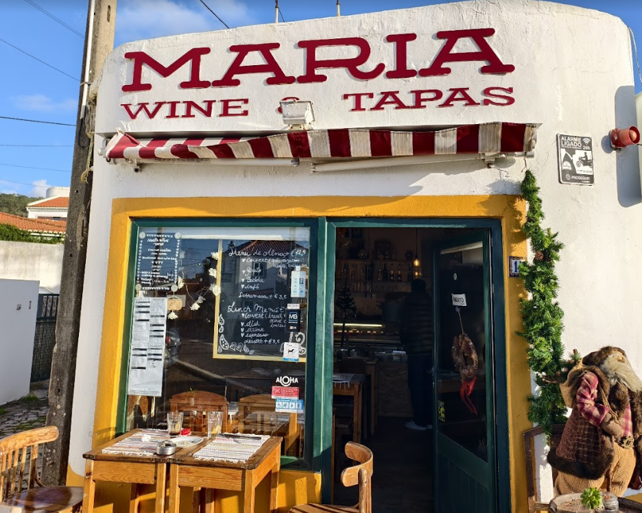 Fachada de um restaurante chamado Maria, que oferece vinhos e tapas, com uma mesa ao ar livre e decoração natalina ao lado da porta.