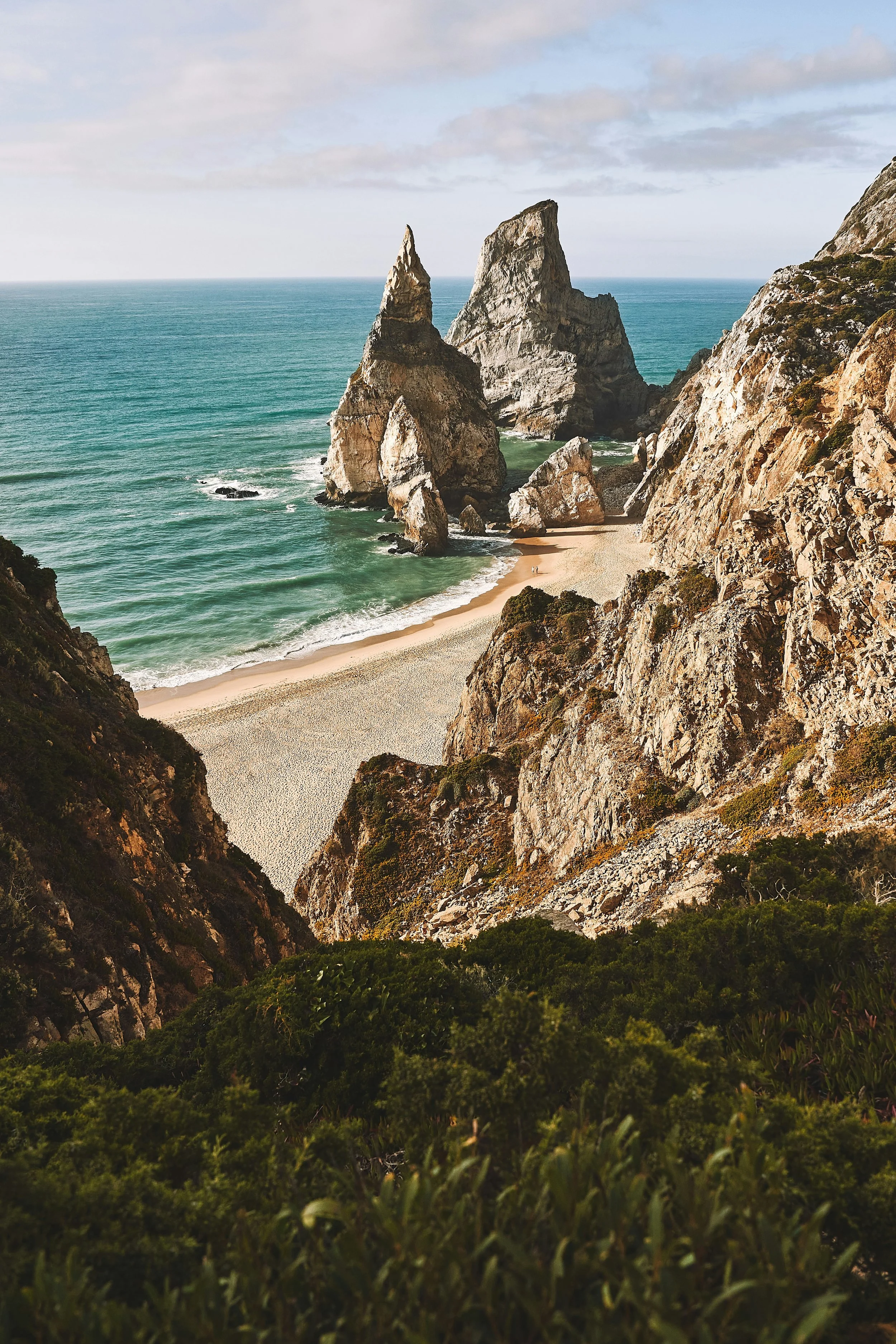 Paisagem costeira com grandes formações rochosas ao mar, uma praia de areia e vegetação verde na margem.