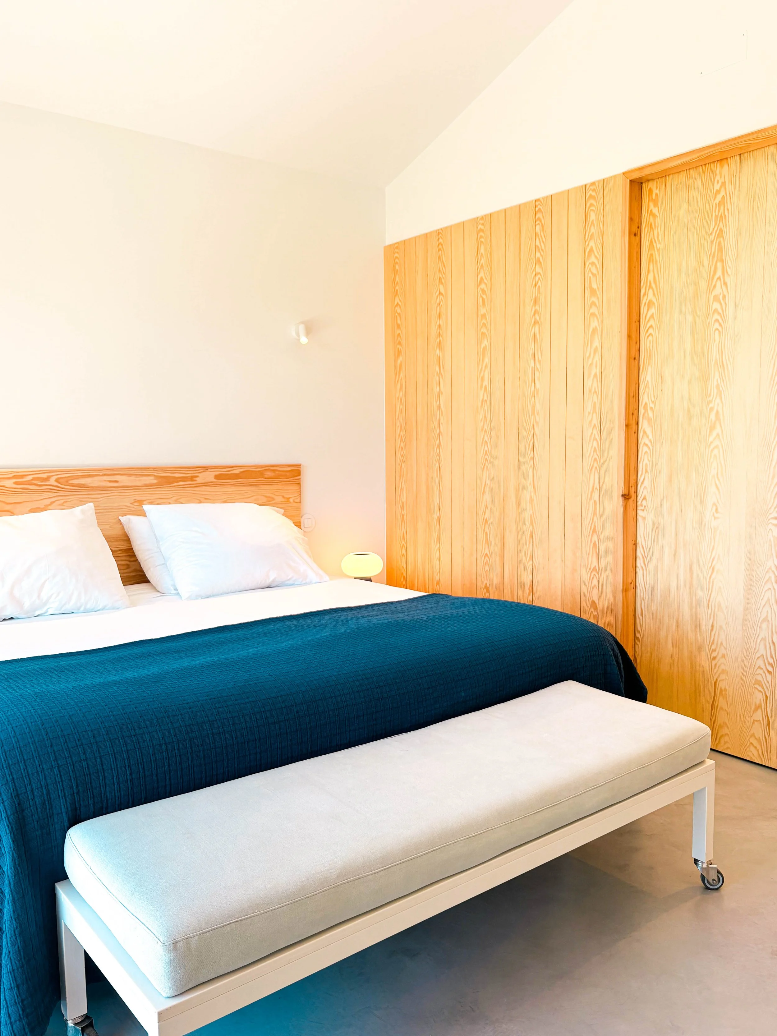 Quarto com cama de casal com cabeceira de madeira, roupa de cama branca e manta azul escura, banco ou chaise longue na frente, paredes brancas e painel de madeira na parede ao fundo, luminária de leitura ao lado da cama.
