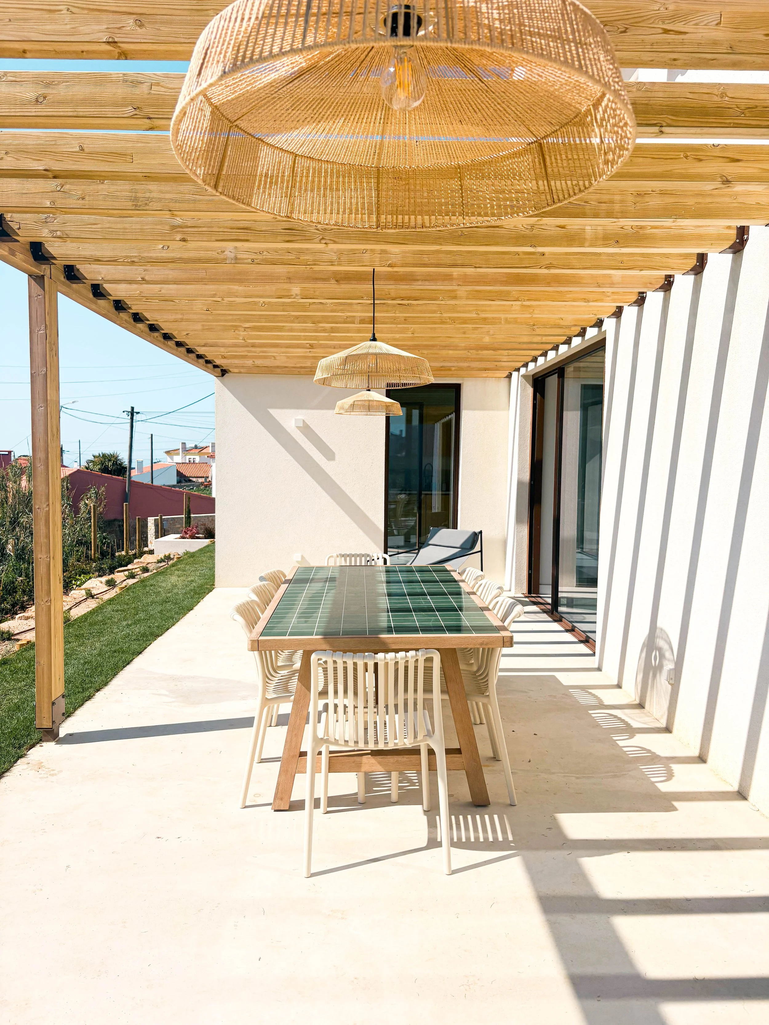 Varanda ao ar livre com mesa de jantar e cadeiras, teto de madeira com pendentes de luz de material natural, sombra projetada por estrutura de vigas, porta de correr de vidro, vista para jardim e casas ao fundo.