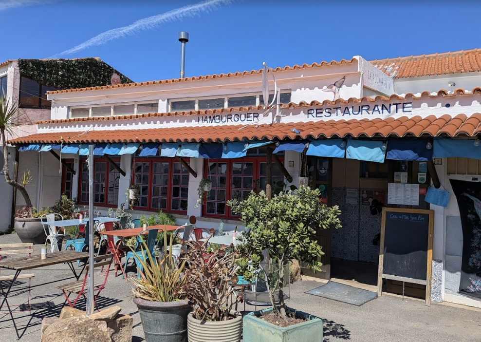 Fachada de um restaurante com mesas ao ar livre, plantas e toldos azuis, com um céu azul ao fundo.