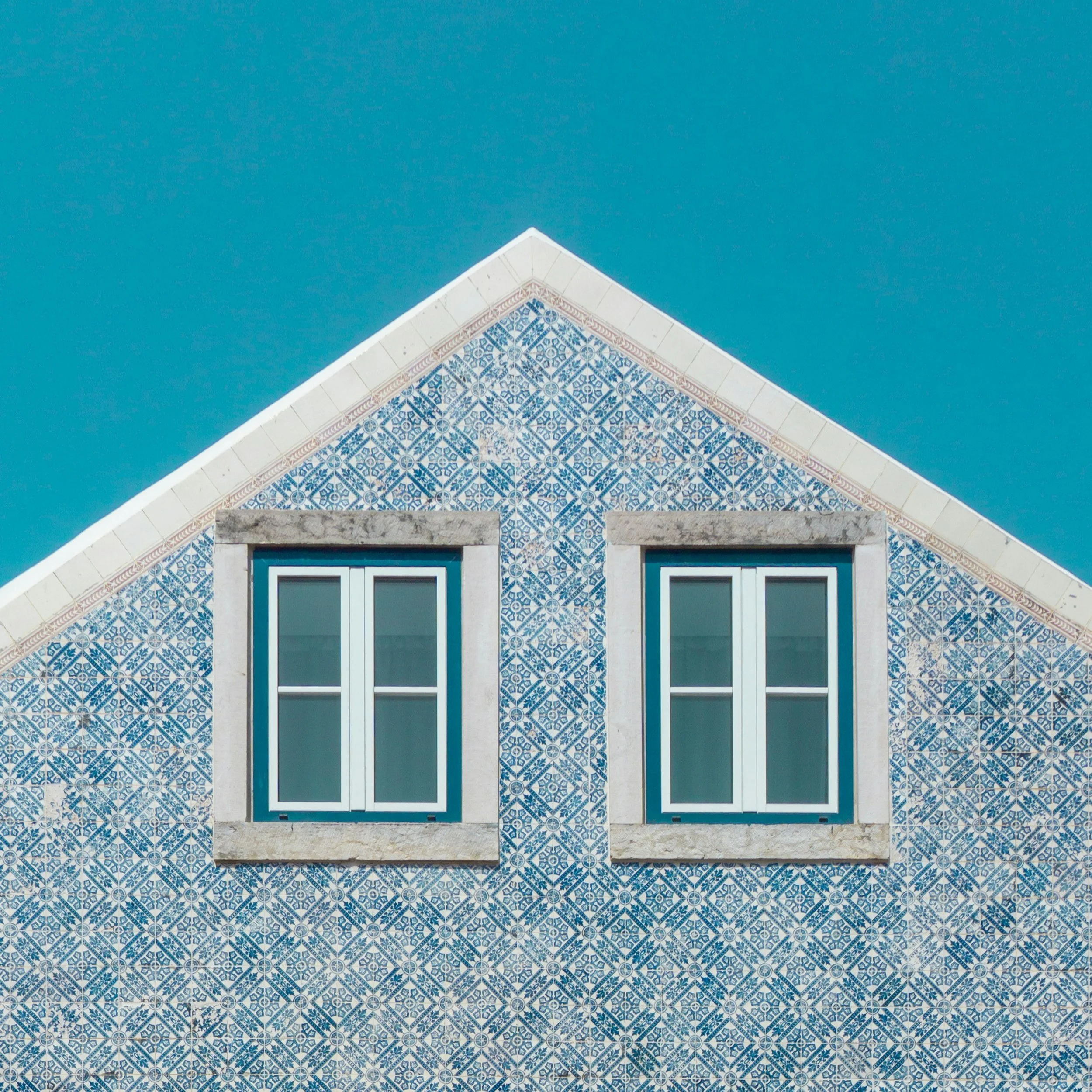 Fachada de uma casa com azulejos azuis e brancos, duas janelas brancas e telhado triangular sob céu azul