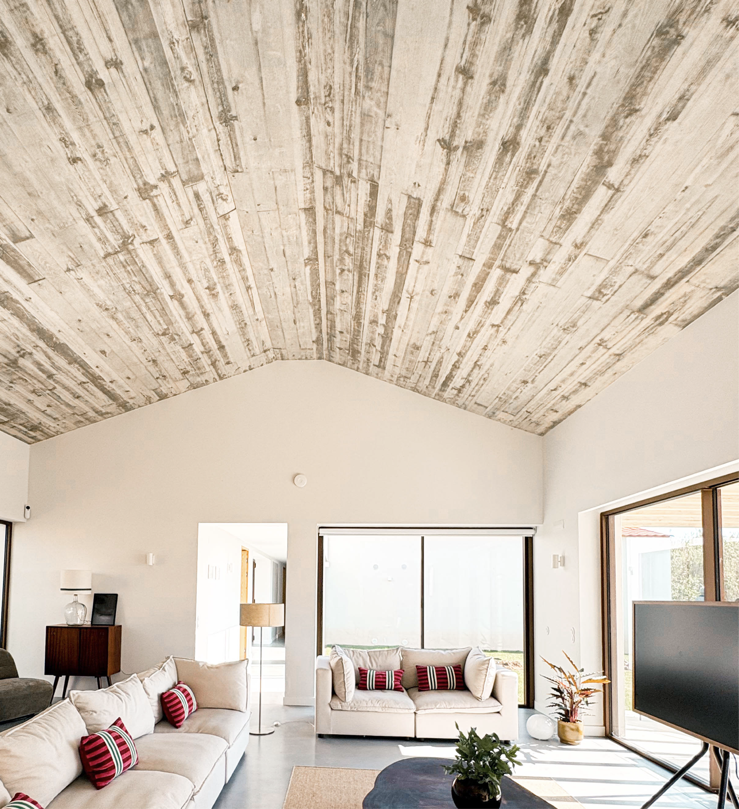 Sala de estar com teto de madeira em estilo rustico, paredes brancas, sofá bege com almofadas vermelhas com listras, janela grande, televisão e planta decorativa.