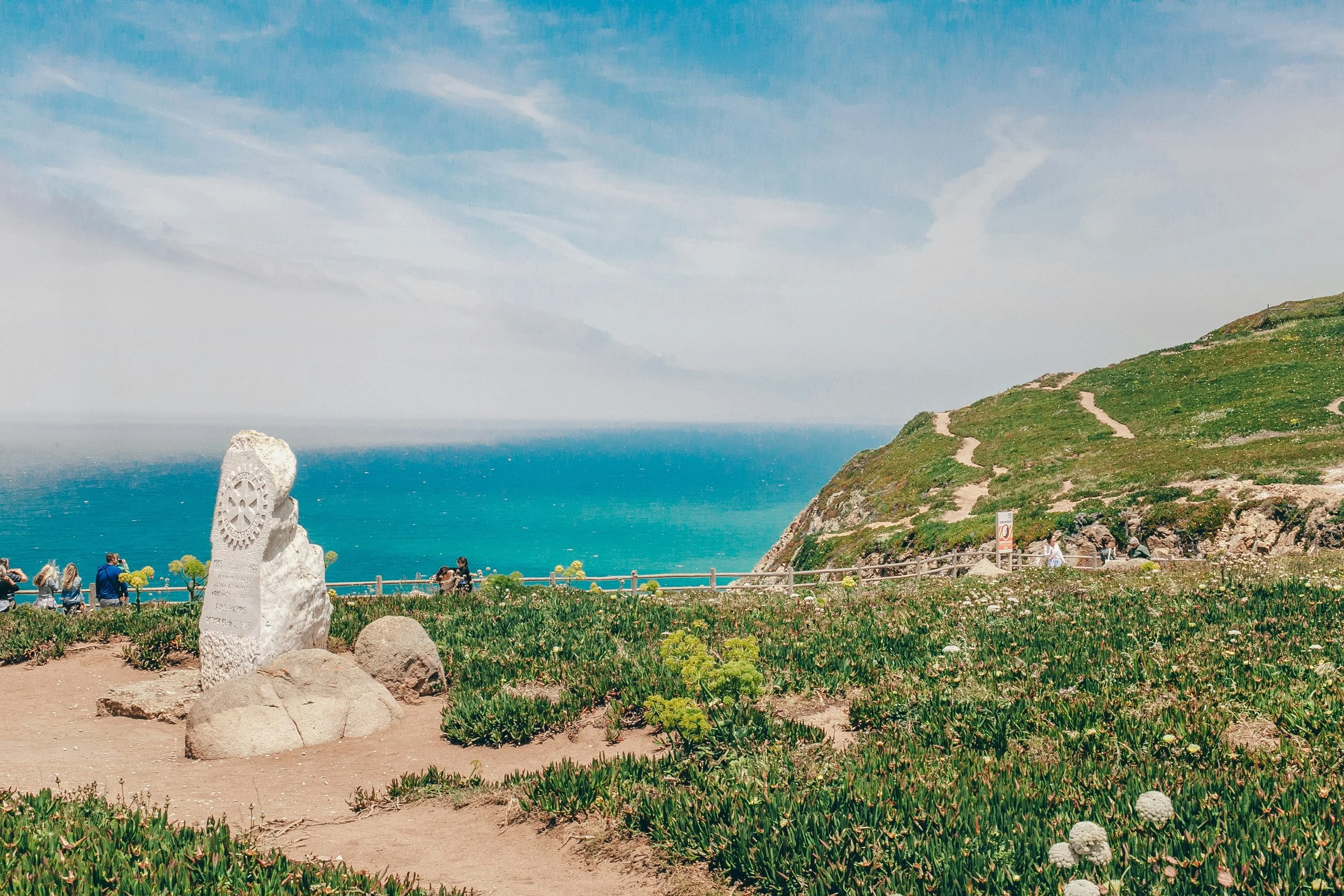 Paisagem costeira com uma vegetação de plantas suculentas, uma escultura de pedra com um símbolo de engrenagem e várias pessoas olhando para o oceano ao fundo, com uma encosta verde e um céu azul com nuvens