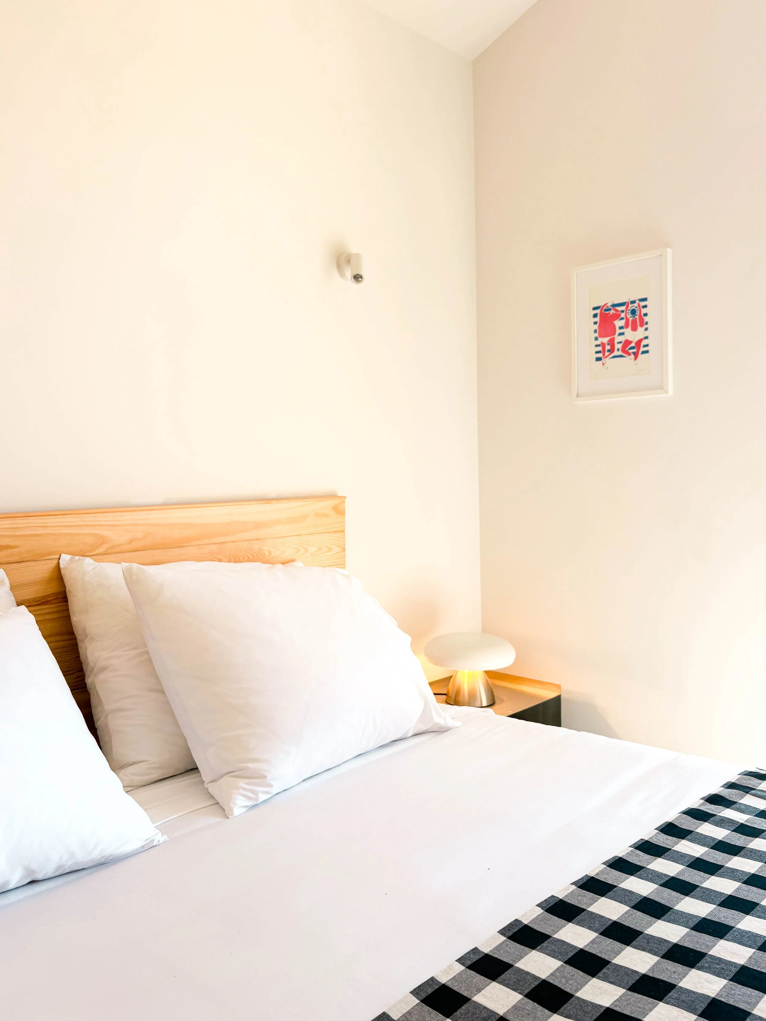 Quarto de hotel com cama de madeira, roupas de cama brancas e almofadas, mesa de cabeceira com luminária moderna, parede branca com quadro decorativo e câmera de segurança na parede.