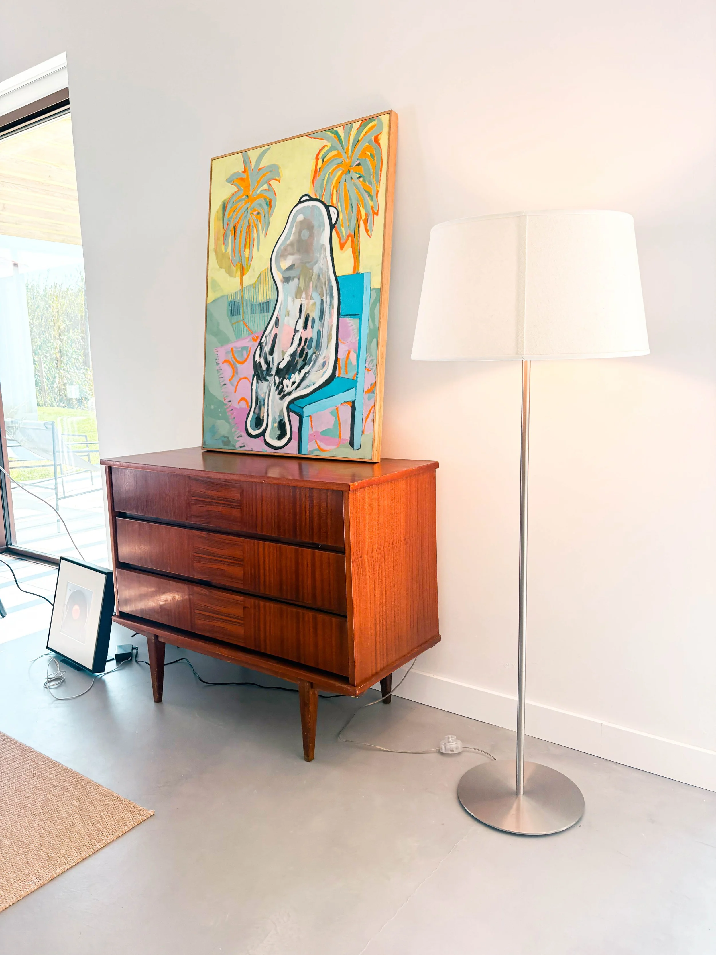 Quadro com arte colorida de um cachimbo sentado em uma cadeira azul, com fundo de árvores palmadas amarelas e laranja, apoiado sobre um móvel de madeira vintage. À direita, luminária de pé. À esquerda, uma porta de vidro que mostra uma área externa e