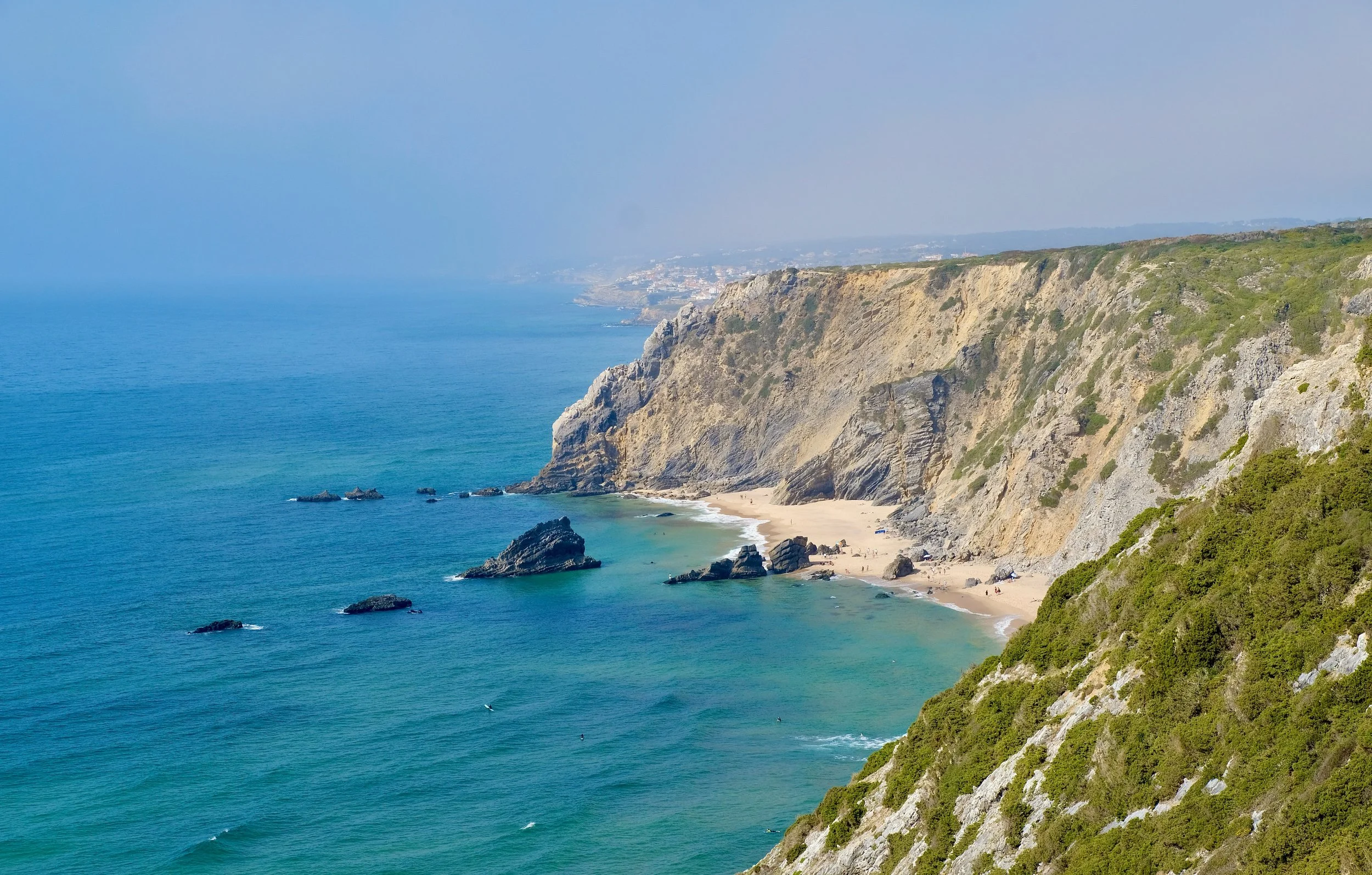 Costa com falésias de vegetação verde e uma praia de areia ao fundo com rochas no mar