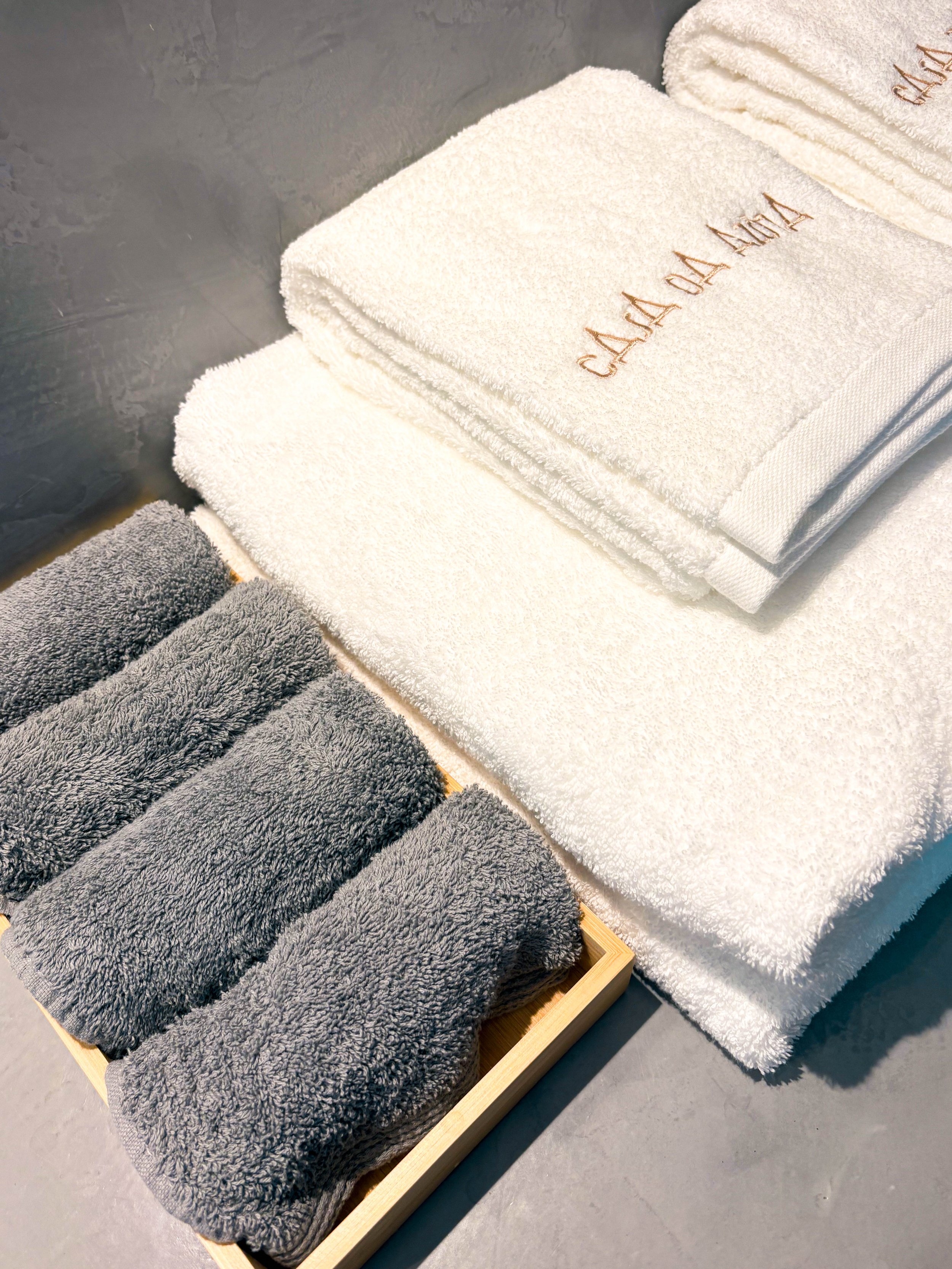 Toalhas brancos e cinzas dobrados sobre uma superfície, com uma caixa de madeira contendo cinco toalhas cinzas. O conjunto parece ser de toalhas de banho ou de rosto, em um ambiente de banheiro ou área de spa.