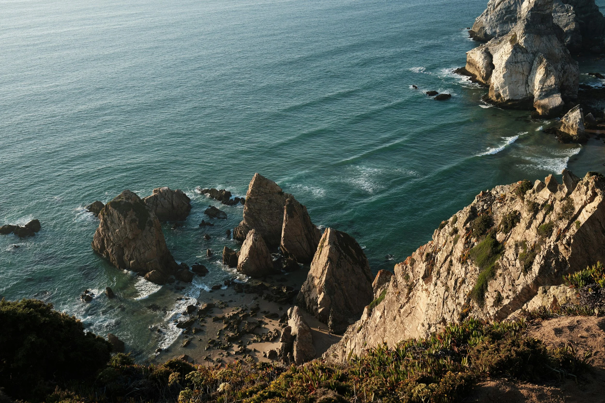 Vista aérea de uma costa rochosa com água do mar azul e verde, com ondas quebrando e formação de rochas e falésias ao longo da costa.
