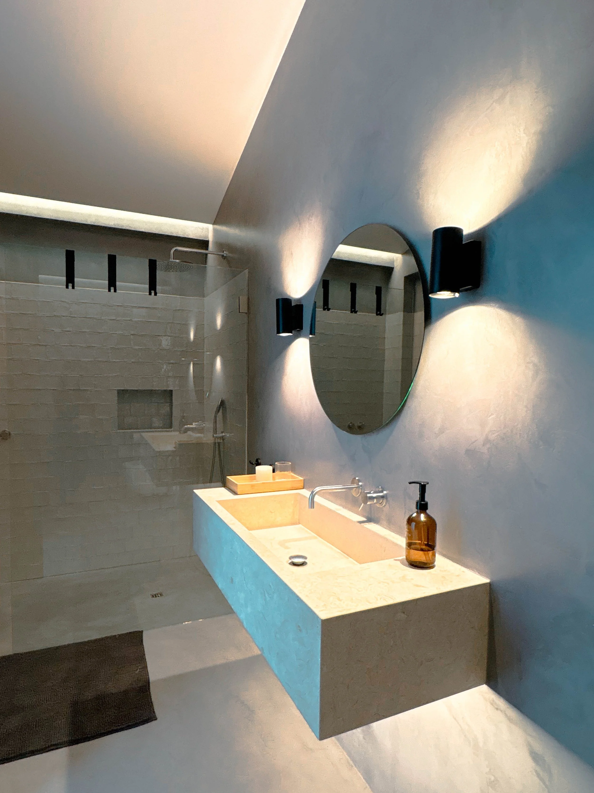 Banheiro moderno com parede de azulejos branco, espelho arredondado, lavabo de pedra, luzes de parede ao redor do espelho, pia com dispenser de sabonete, área de banho com ducha e teto inclinado.