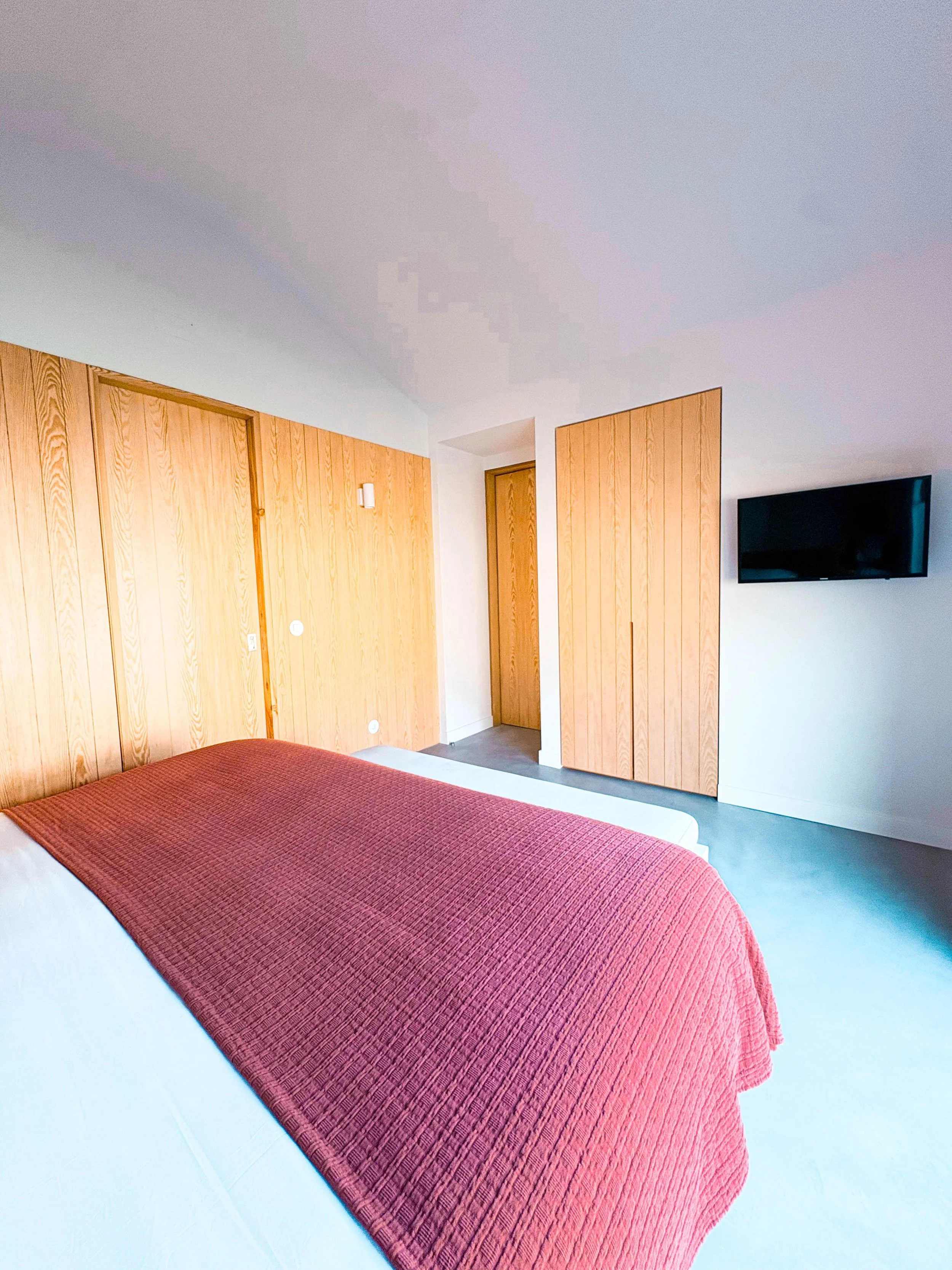 Quarto com cama de roupa branca e cobertor vermelho, paredes de madeira clara, porta de madeira na parede e televisor de tela plana na parede branca.