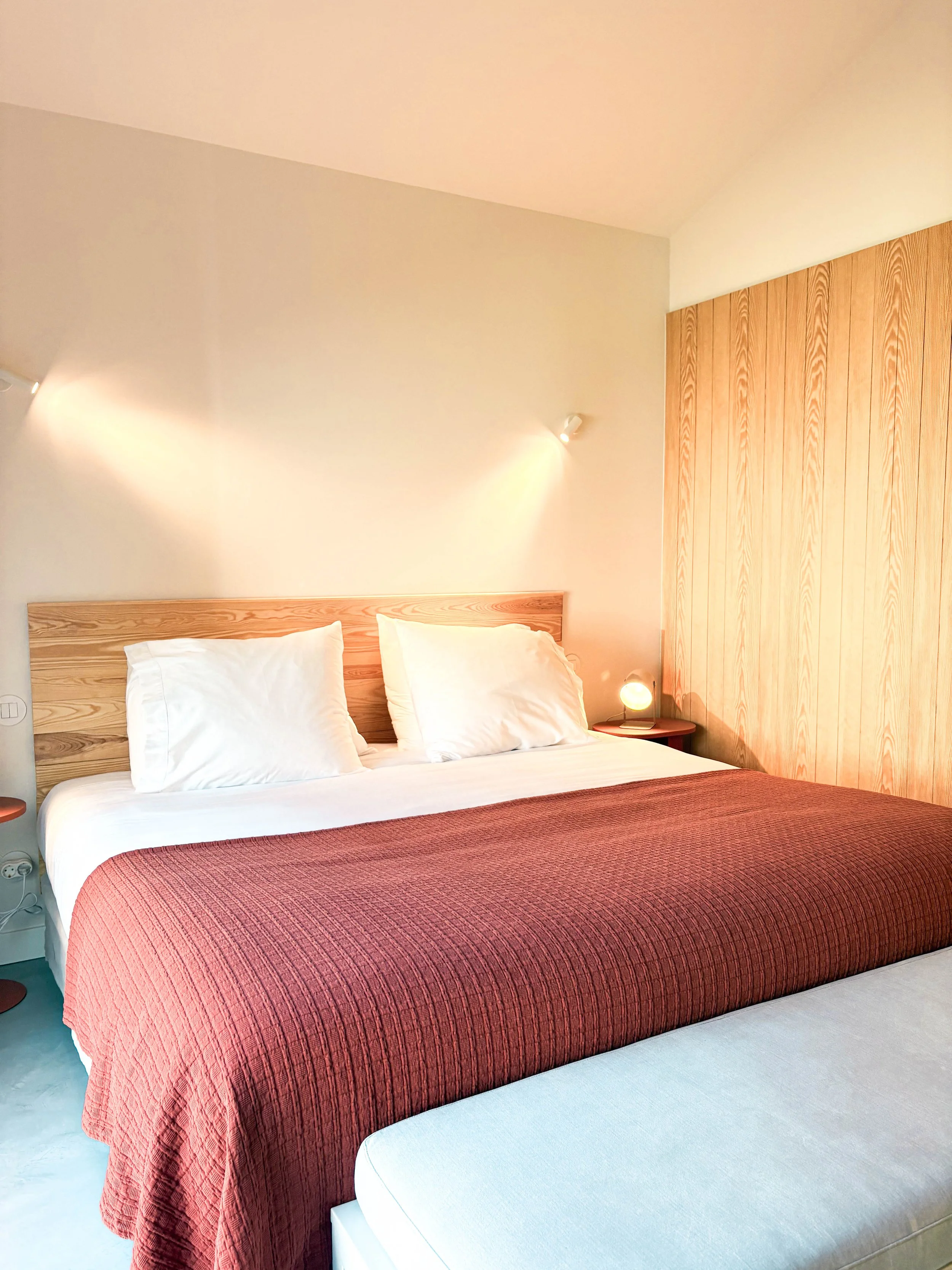 Quarto de hotel com cama de casal com duas almofadas brancas, cabeceira de madeira, edredom vermelho, parede de madeira ao lado e iluminação suave