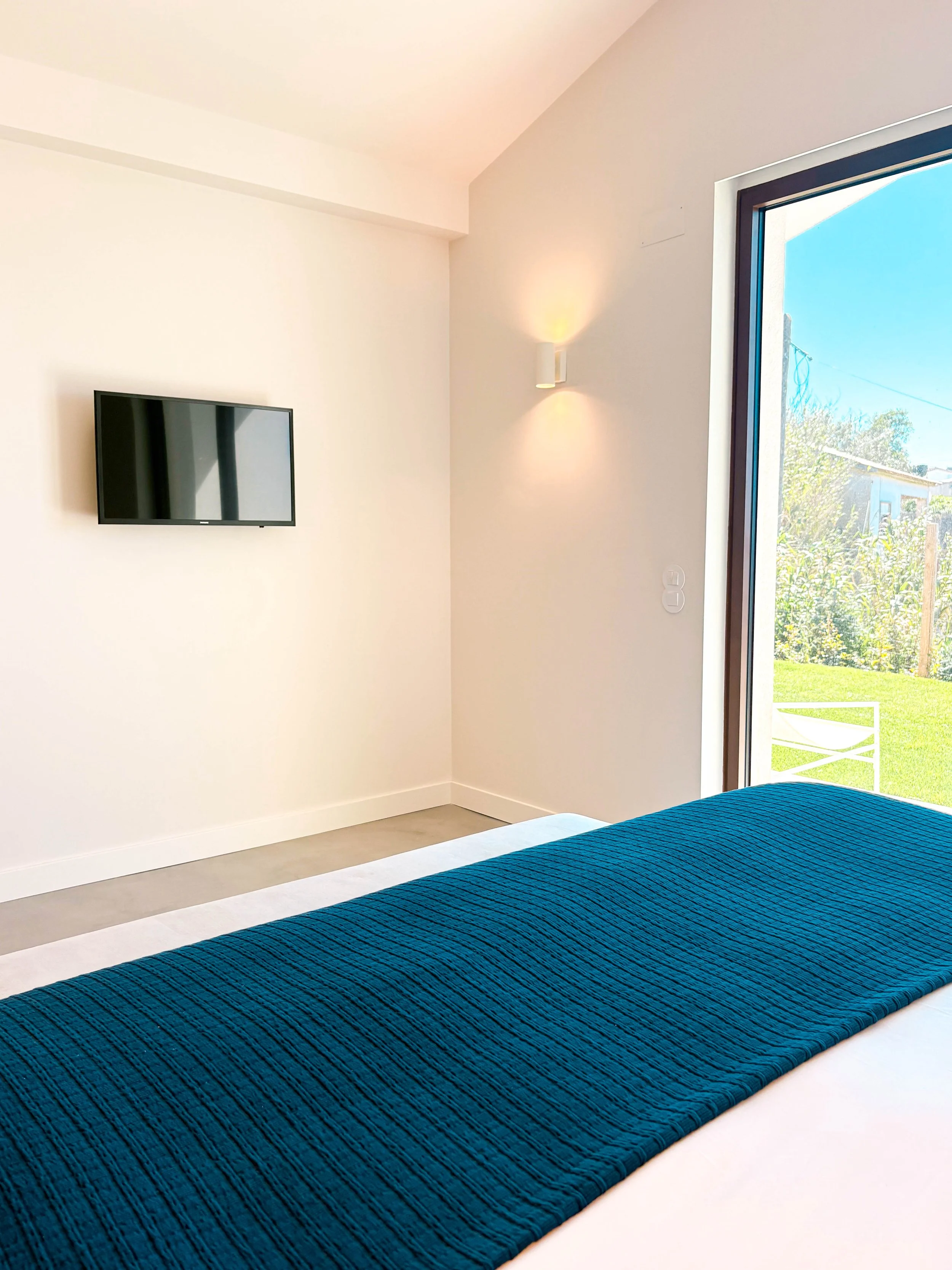 Dormitório com janelas amplas, parede branca, televisão na parede, luminária de parede, roupa de cama branca com colcha azul, vista do jardim externo.