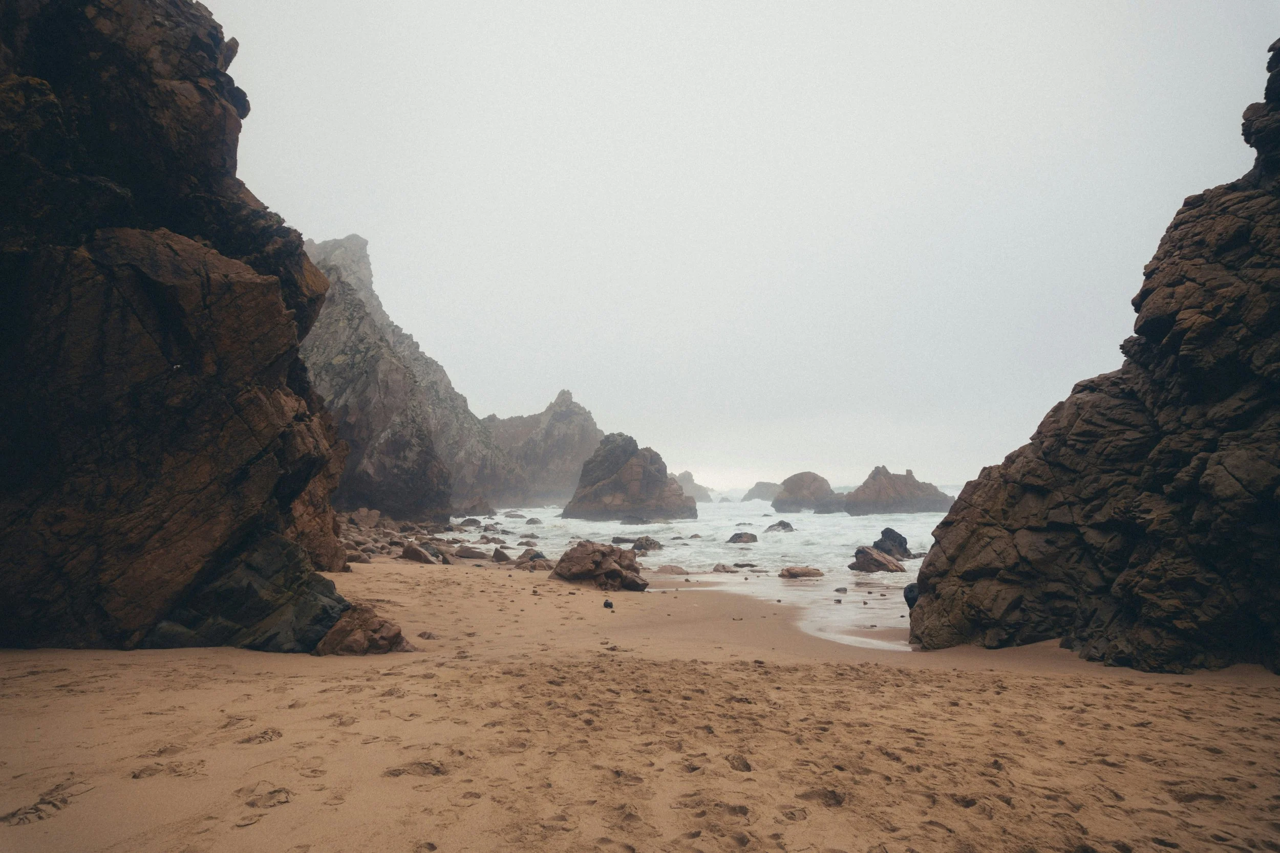 Praia com areia, rochas grandes nas margens e no mar, céu nublado e bruma ao horizonte.