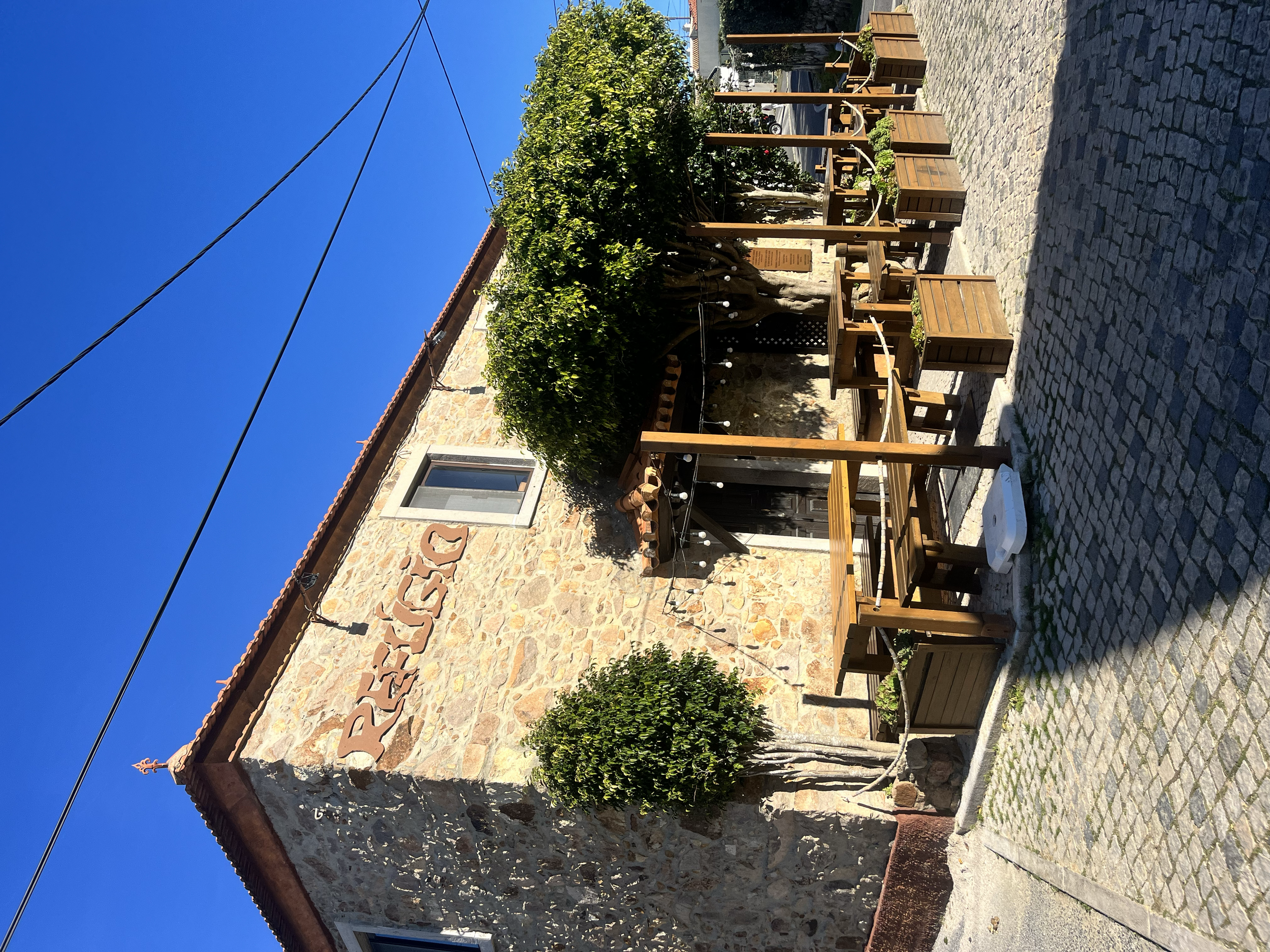 Fachada de uma casa de pedra com o nome 'PRESUCO' em destaque. Área externa com mesas de madeira e árvores decorativas, sob céu azul claro.