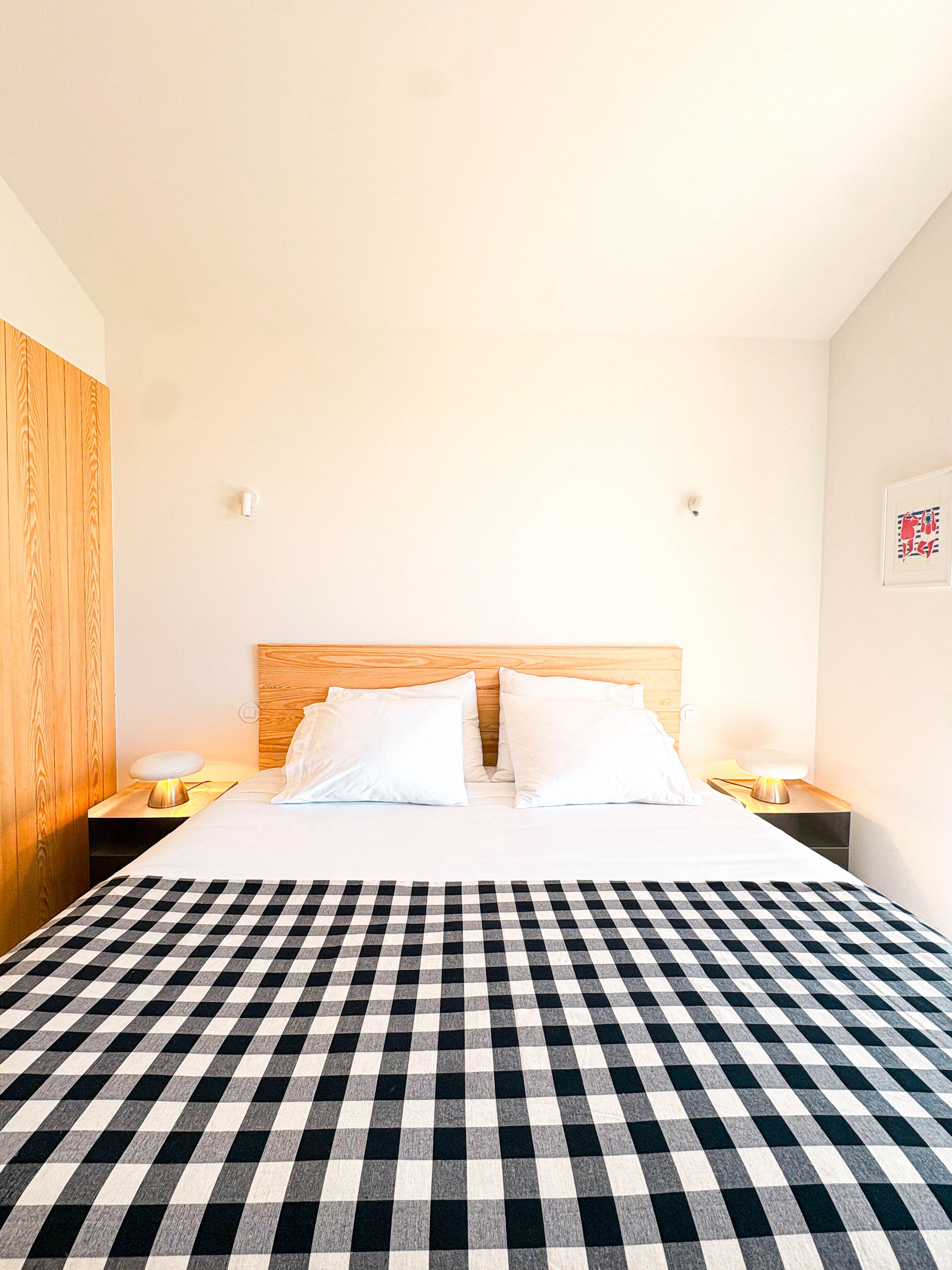 Cama de casal com cabeceira de madeira, roupa de cama branca, travesseiros brancos e um cobertor de tecido listrado em preto e branco, em um quarto com paredes brancas e iluminação suave.