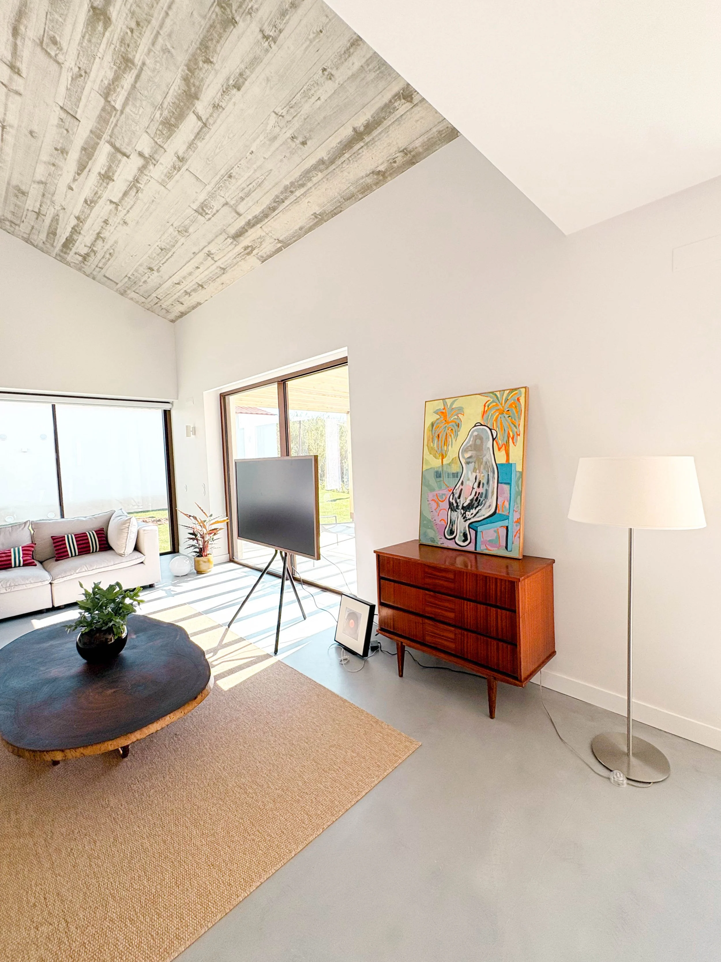 Sala de estar moderna com sofá branco, mesa de centro de madeira escura, tapete bege, televisão em suporte, pintura colorida ao lado de um móvel de madeira, luminária de piso branca, janelas grandes com vista para o exterior.