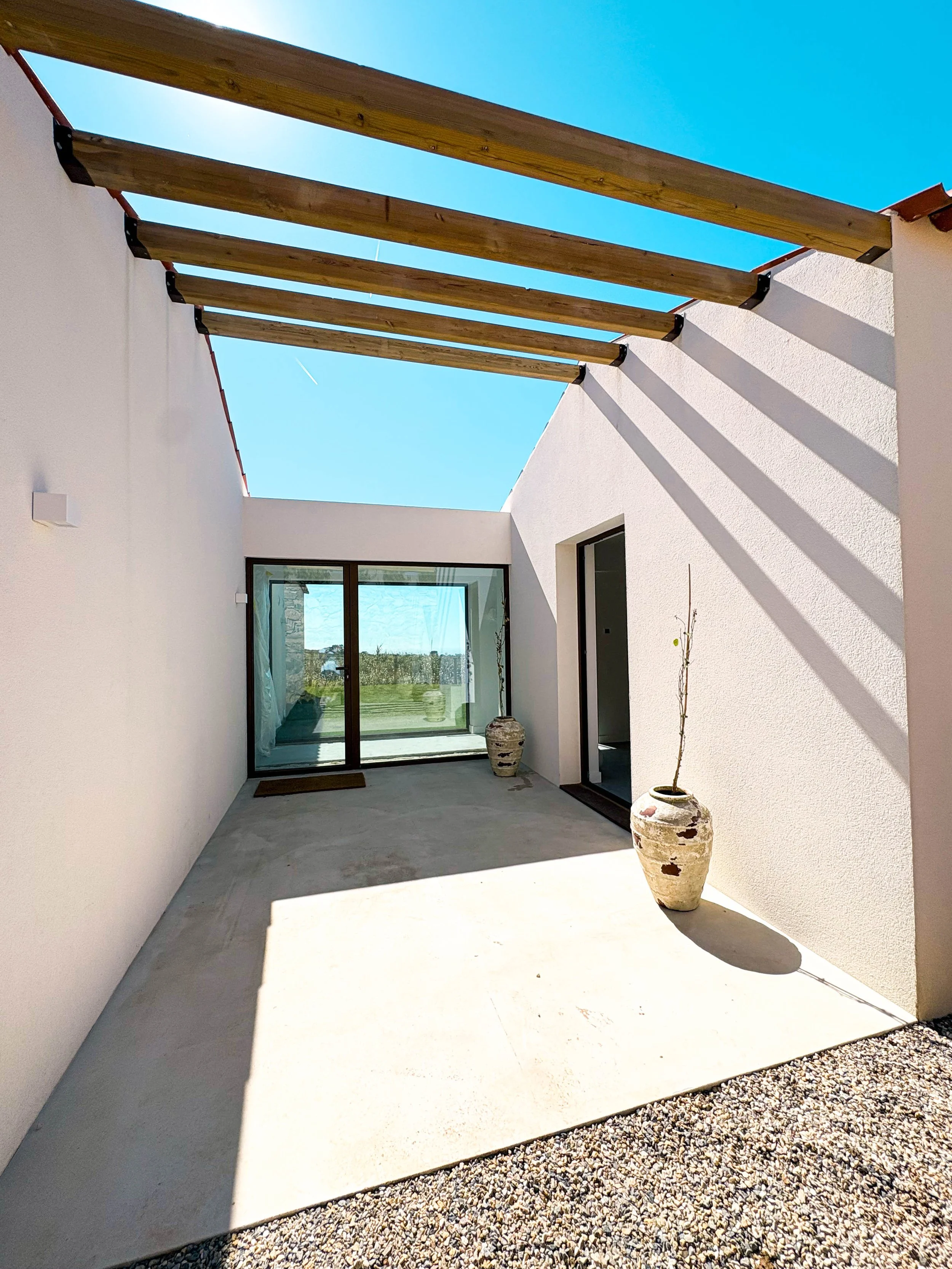 Varanda de casa moderna com paredes brancas, piso de cimento, dois vasos de cerâmica com plantas, uma porta de vidro com cortina e uma estrutura de madeira sem cobertura.