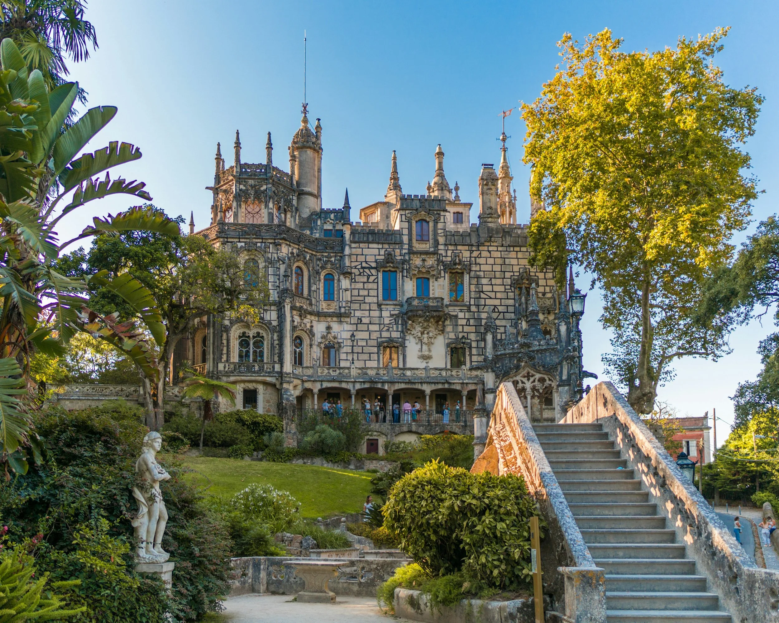 Castelo de Pena em Sintra, Portugal, com escada de pedra na frente, jardim com estátua de uma criança, árvores verdes e céu azul ao fundo.