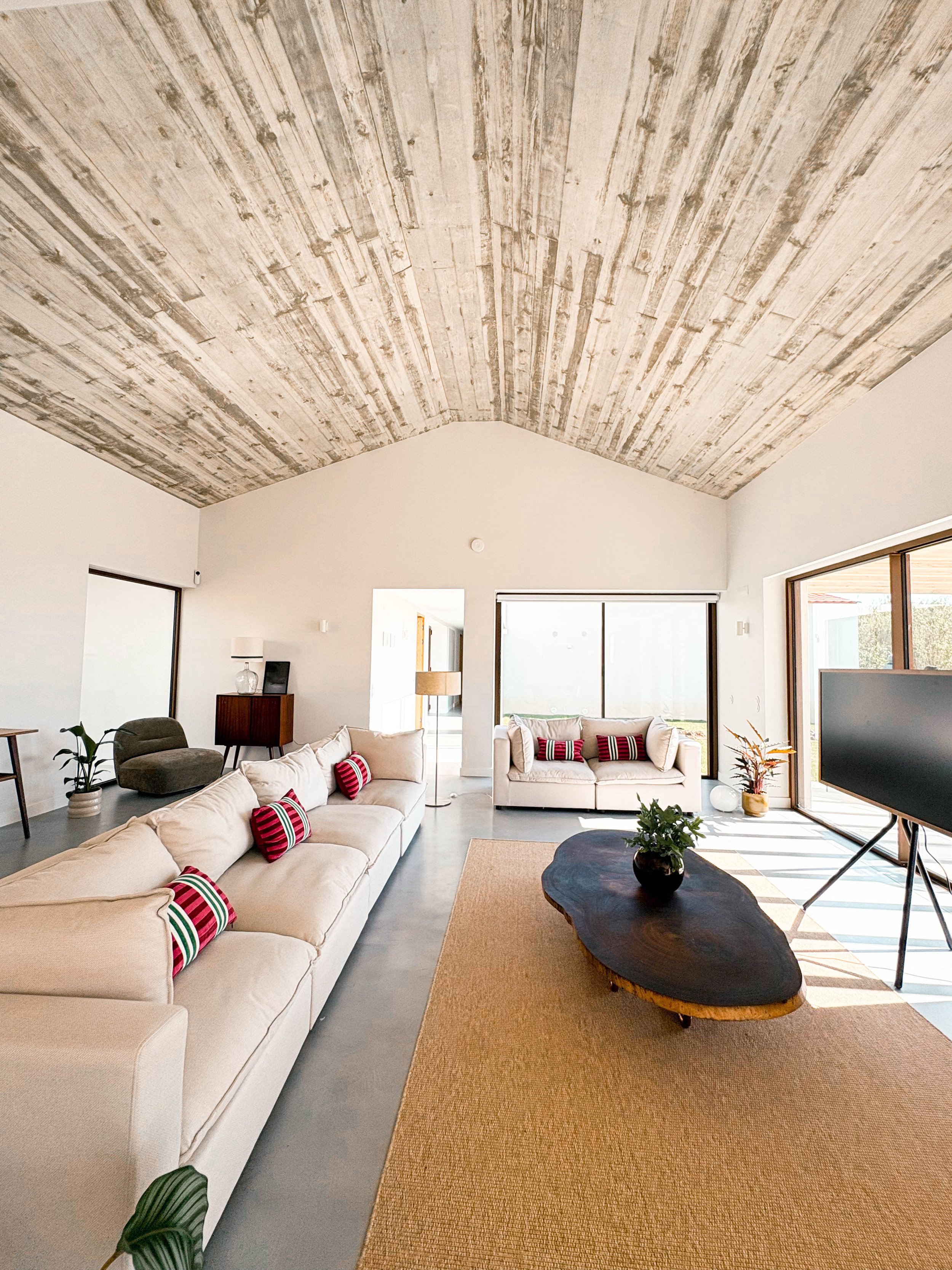 Sala de estar com teto de madeira, sofá claro com almofadas vermelhas, mesa de centro de madeira escura, televisão em tripé, janela grande com vista para o exterior, detalhes decorativos e plantas