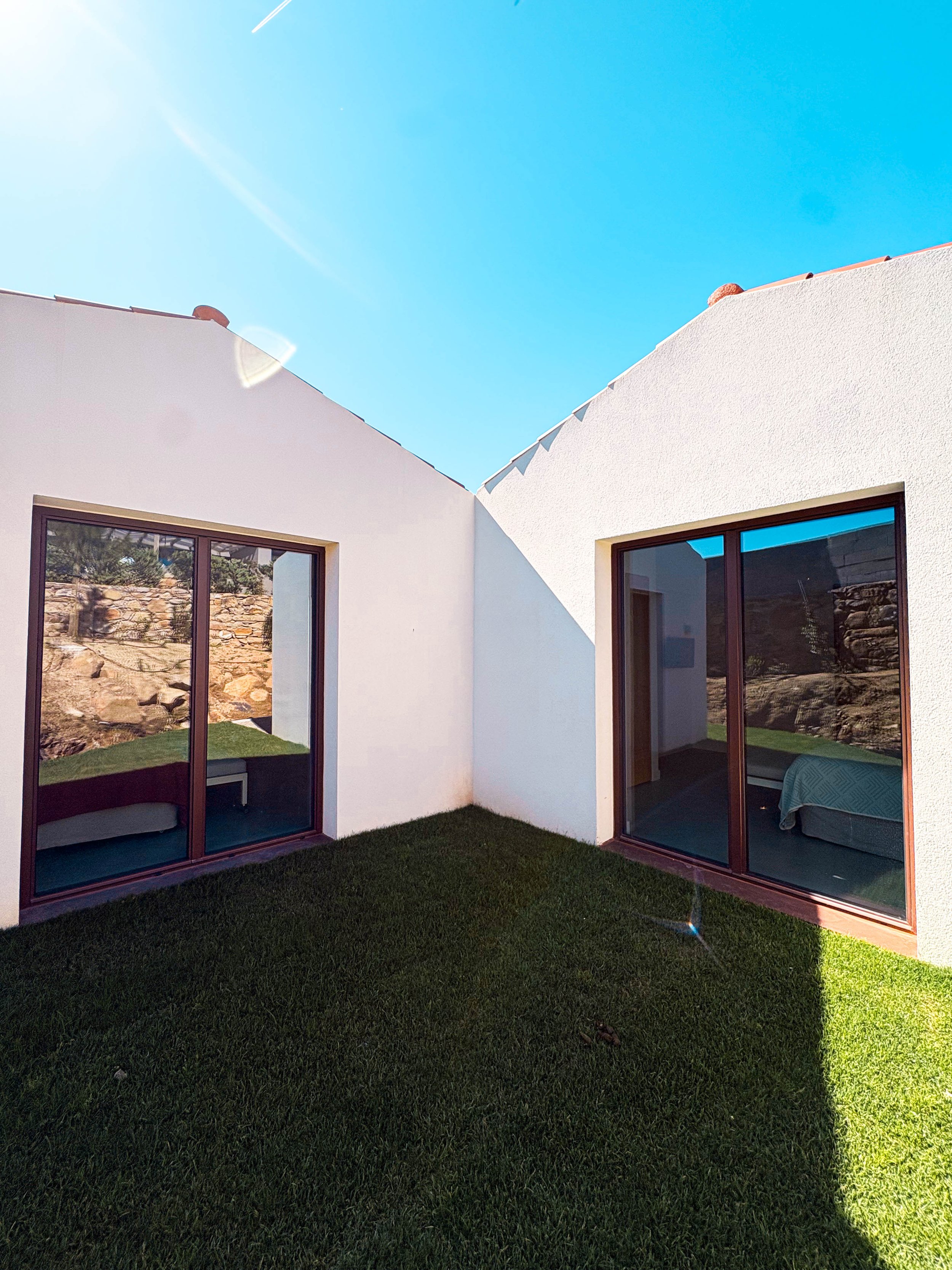 Jardim com gramado em frente a janelas de vidro de duas casas com paredes brancas