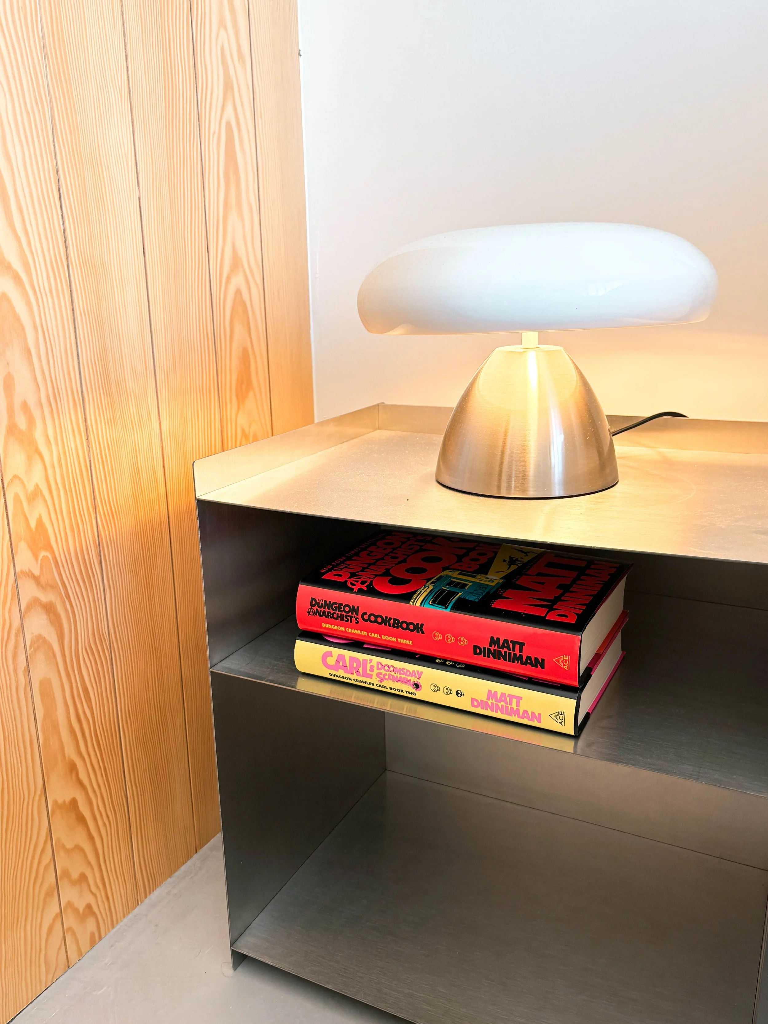 Lâmpada moderna de abajur de metal dourado e luz branca aquecida, colocada sobre uma mesa de canto de metal escuro. Ao lado, há dois livros empilhados com capas coloridas, um em preto e vermelho ('Dungeon...') e outro em amarelo e rosa ('Carl's Dooms