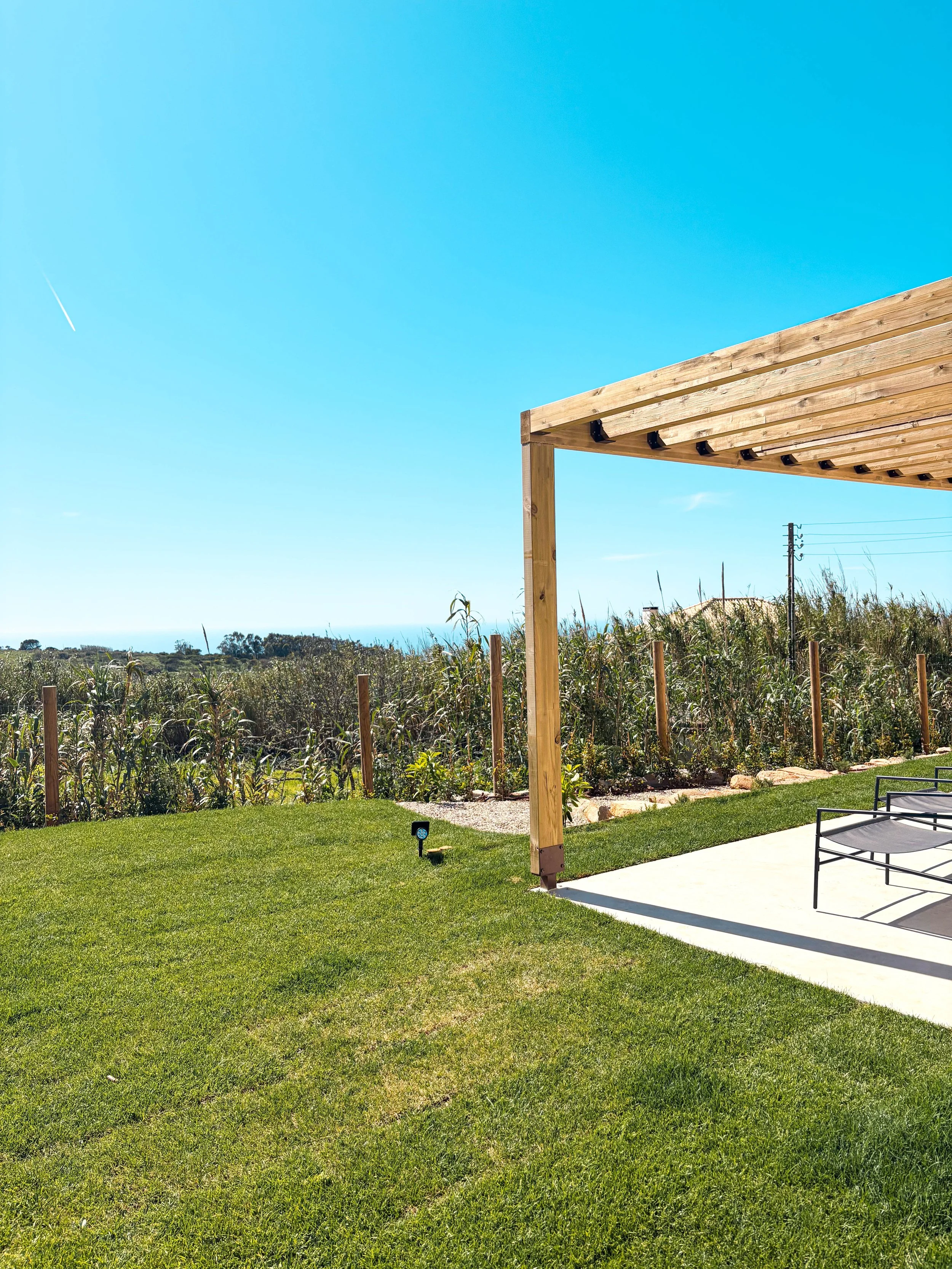 Jardim com gramado bem cuidado, cerca de madeira, uma estrutura de cobertura de madeira, mobiliário de quintal e vegetação ao fundo, sob céu azul claro.