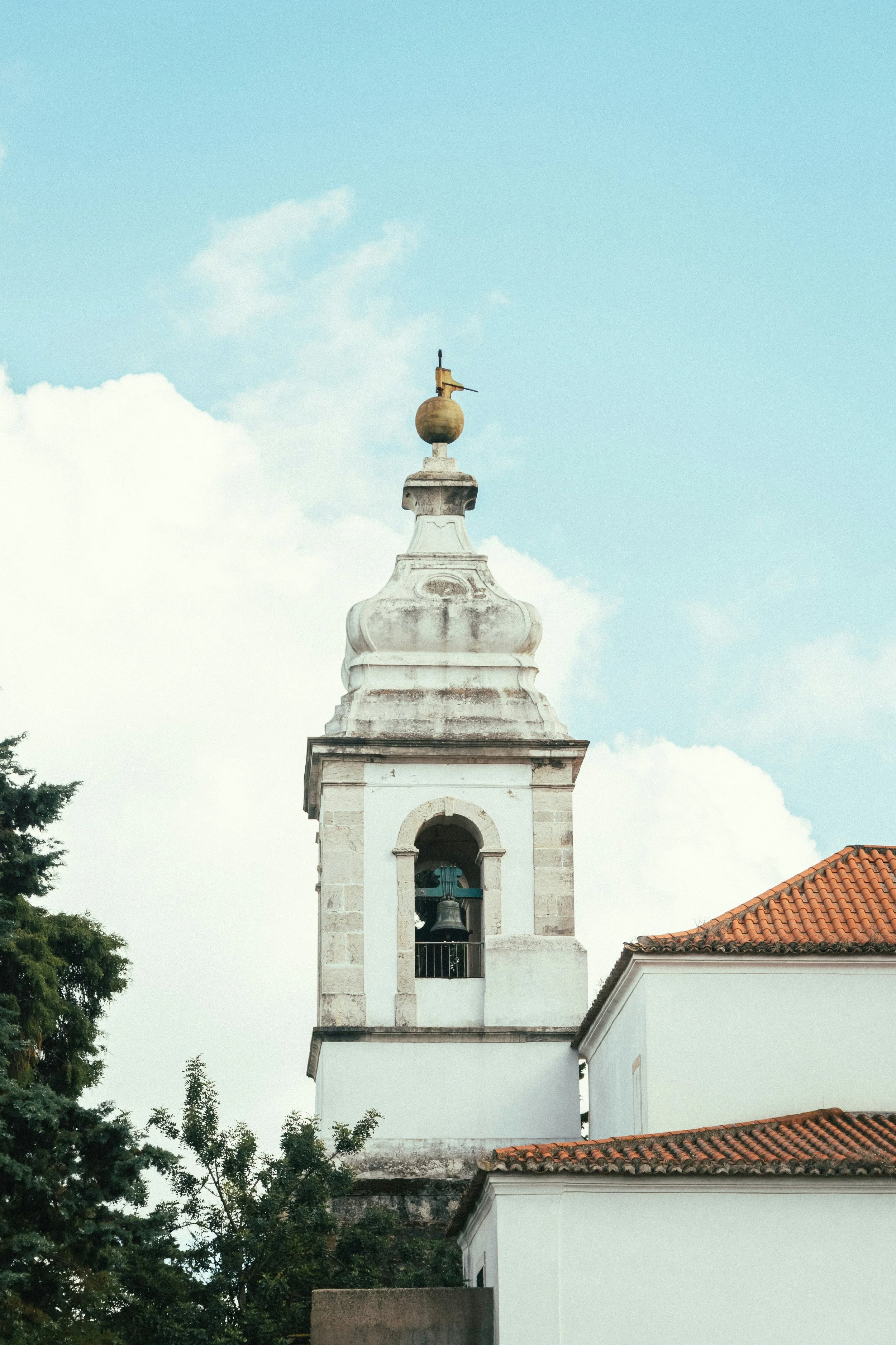 Igreja colonial com torre e sino, telhado de telhas cerâmicas, cercada por árvores, céu com nuvens.