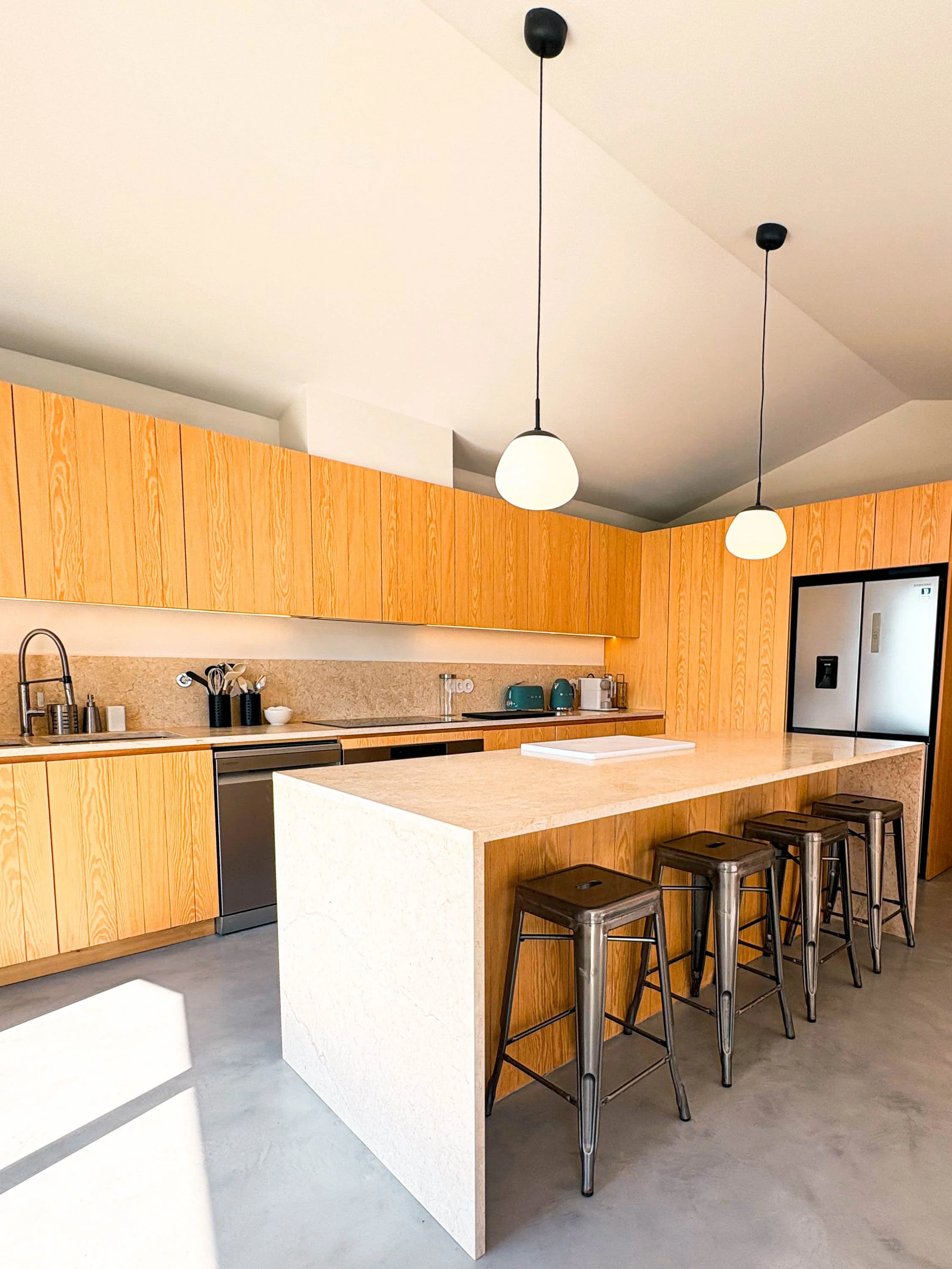 Cozinha moderna com armários de madeira clara, ilha central de pedra branca, quatro banquetas metálicas e iluminação com dois globos brancos pendurados no teto.