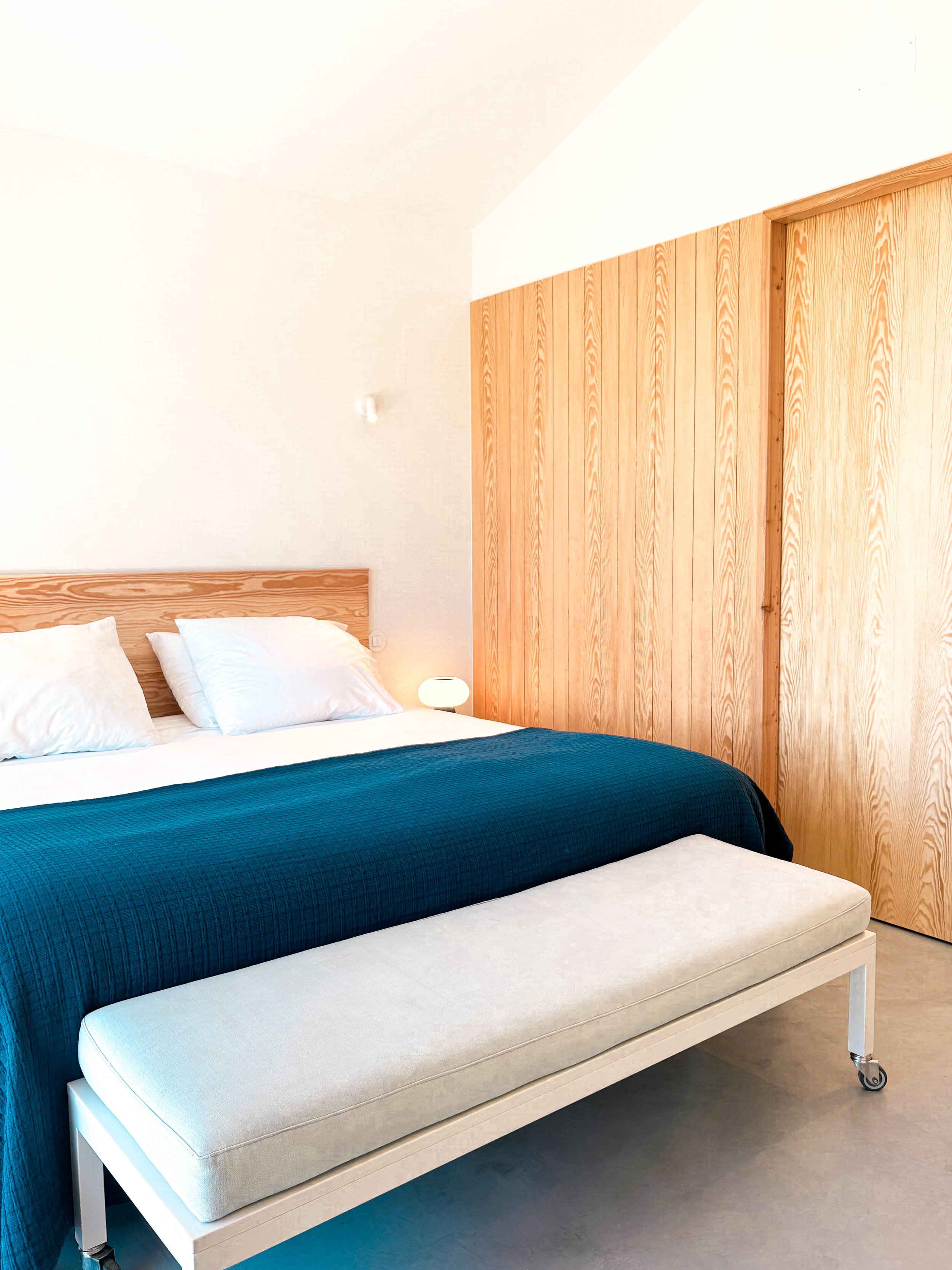 Quarto de hotel com cama de casal, roupas de cama brancas, travesseiros brancos, edredom azul escuro, banco cinza, parede de madeira ao lado da cama, parede branca, iluminação suave.