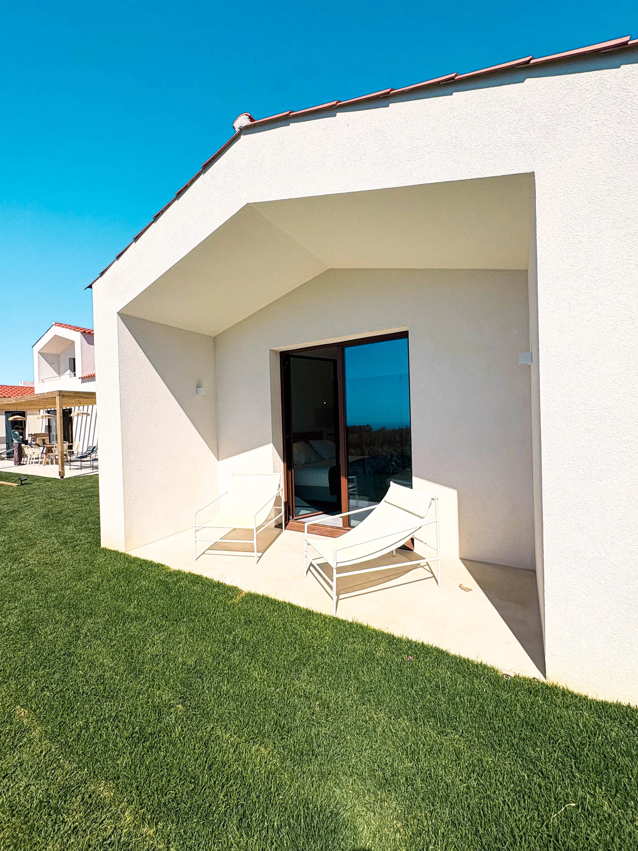 Residência moderna com varanda, duas cadeiras brancas, grama bem cuidado e céu azul.