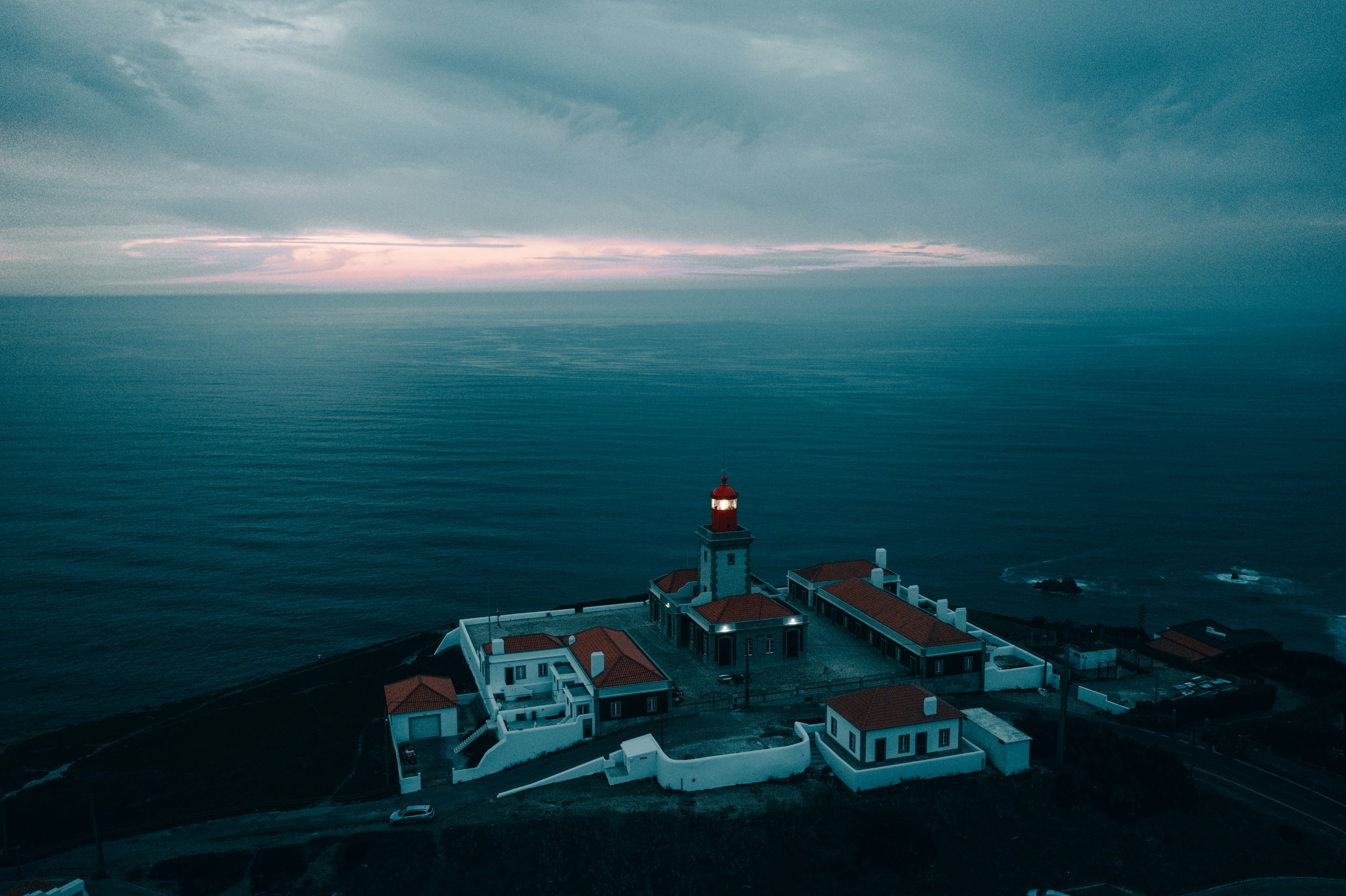 Faro com luz vermelha em uma ponta de uma ilha cercada pelo oceano, sob céu nublado ao entardecer.