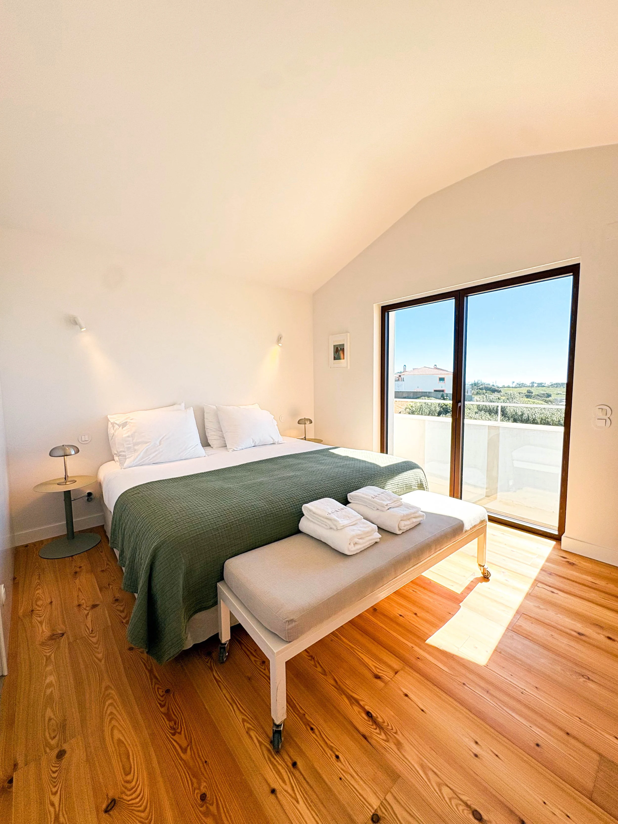 Quarto de hotel com cama de casal, janelas grandes com vista para o exterior, móveis simples e decoração minimalista.