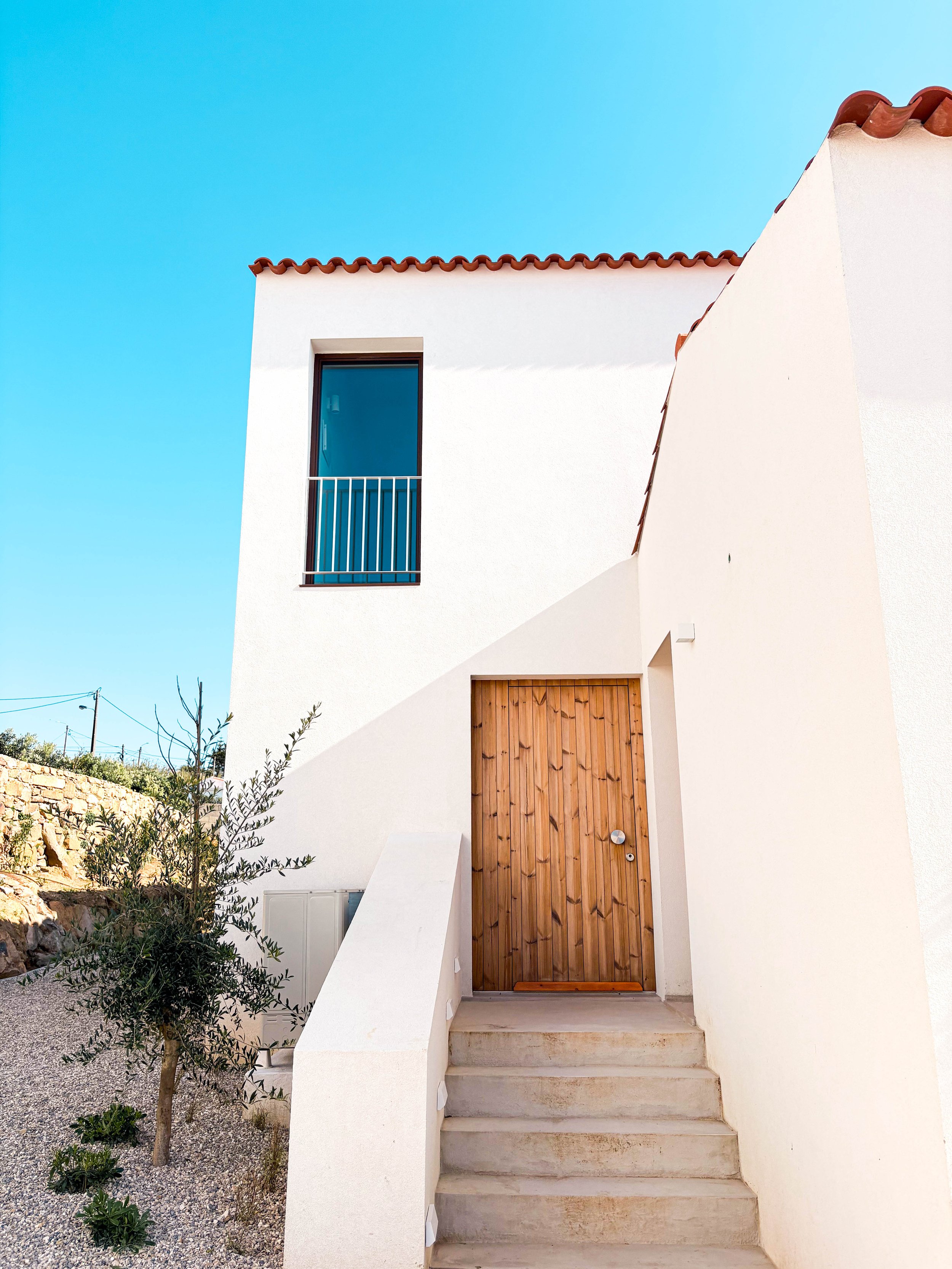 Fachada de uma casa moderna com parede branca, porta de madeira, escada de cimento, janela com grades e jardim com árvore pequeno, sob céu azul.